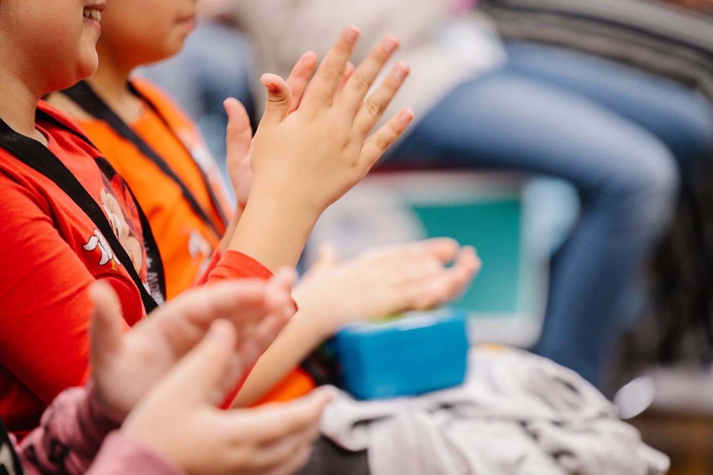 Kinder sitzen und klatschen in die Hände, sie tragen bunte Kleidung. Der Fokus liegt auf ihren Händen, der unscharfe Hintergrund zeigt andere sitzende Personen.