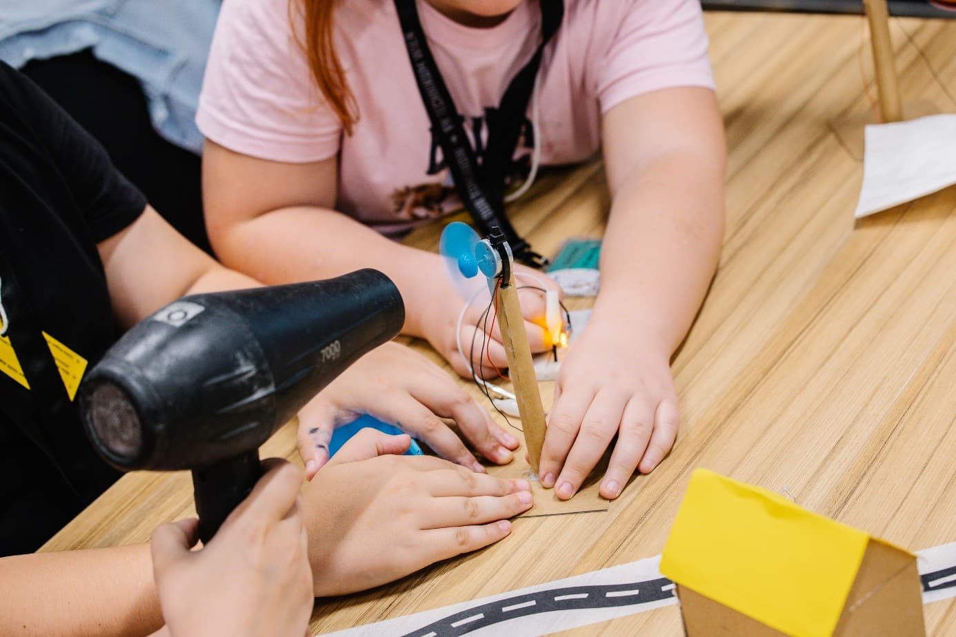 Drei Kinder arbeiten gemeinsam an einem kleinen windbetriebenen Projekt an einem Tisch; eines hält einen Haartrockner, der Luft auf einen blauen Ventilator bläst, während die anderen das Projekt, das Teil einer praktischen MINT-Aktivität ist, überwachen.