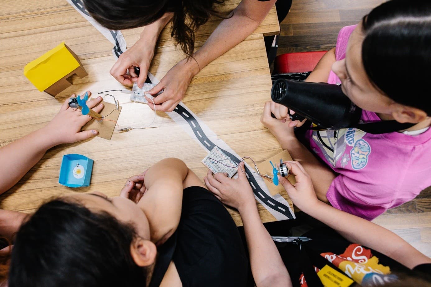 Kinder und ein Erwachsener arbeiten gemeinsam an einem Tisch an einem naturwissenschaftlichen oder technischen Projekt, indem sie kleine elektronische Bauteile und Kartonteile entlang einer Papierstraße zusammensetzen. Die Ansicht ist von oben und zeigt aktive Hände und konzentrierte Gesichter.