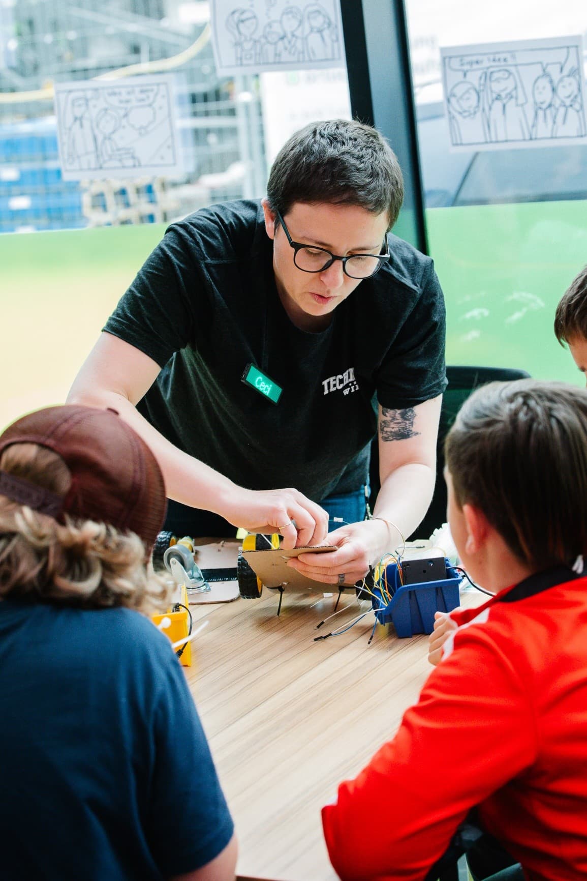 Eine Person mit kurzen Haaren und Brille hilft drei Kindern beim Zusammenbau eines kleinen Roboters an einem Tisch. Elektronische Bauteile liegen verstreut auf dem Tisch. An ein Fenster im Hintergrund sind Zeichnungen geklebt.