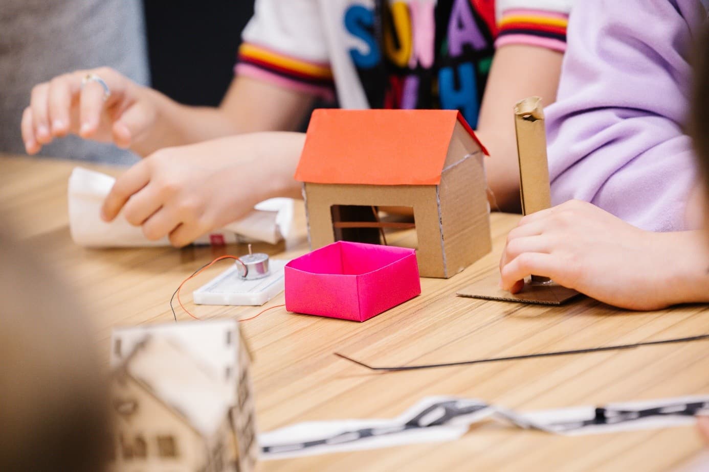 Kinderhände, die an einem Tisch an einem Bastelprojekt arbeiten und kleine Kartonmodelle zusammensetzen, darunter ein Haus mit rotem Dach und eine rosa Schachtel.