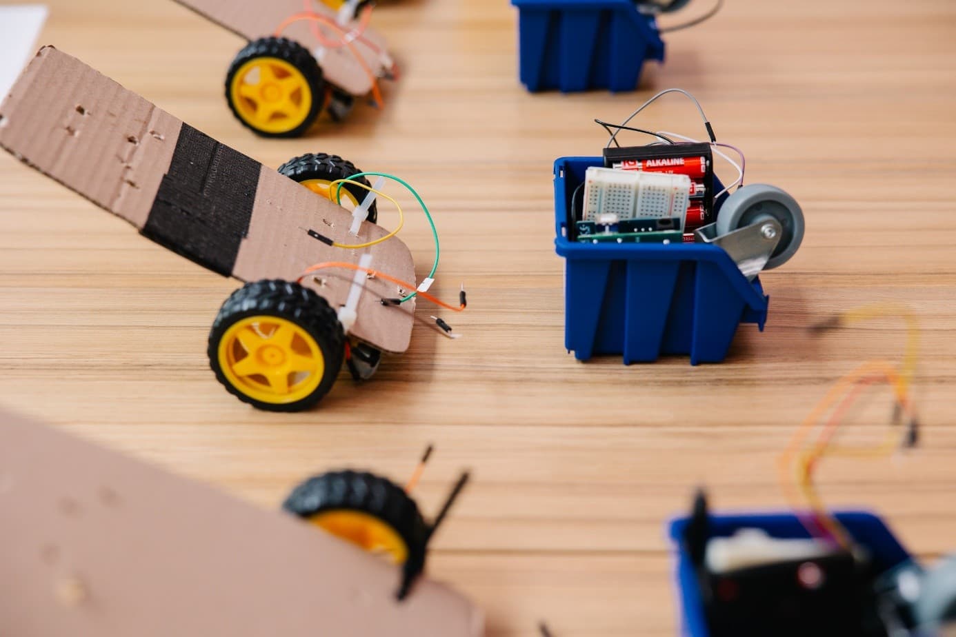 Kleine Roboter auf Rädern aus Pappe, Plastikbehältern und elektronischen Bauteilen sind auf einem Holztisch angeordnet und zeigen DIY-Robotikprojekte.