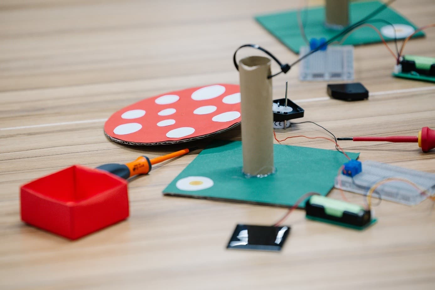 Auf einem Tisch liegt ein Bastelprojekt mit einer Pappröhre, einem roten Kreis mit weißen Punkten, elektronischen Bauteilen, Drähten und einem roten Kasten, die alle auf dünnen grünen Platten angeordnet sind.