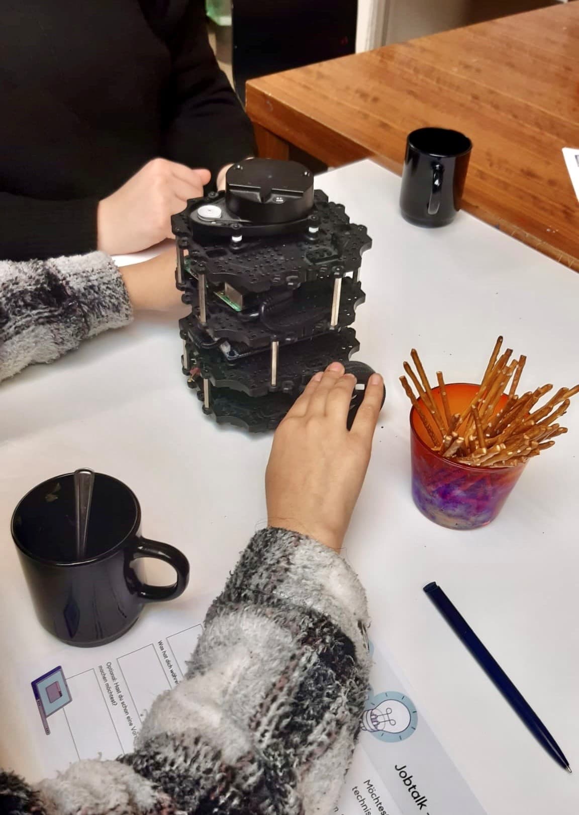 Zwei Personen sitzen an einem Tisch mit einem schwarzen Roboter, einem schwarzen Becher, einer Tasse mit Brezelstangen und Papieren. Eine Person greift nach dem Roboter; ein Stift und ein weiterer Becher sind ebenfalls zu sehen.