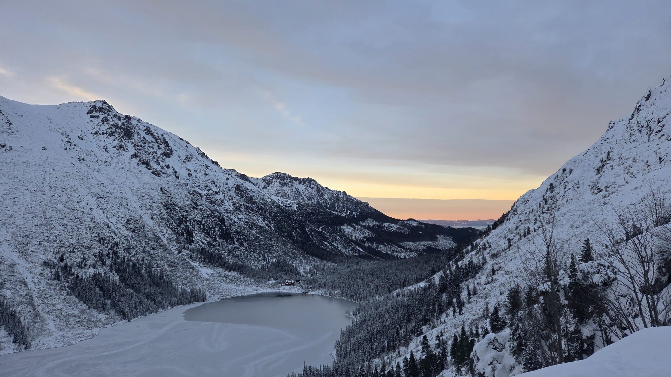 Schneebedeckte Berge umgeben einen teilweise zugefrorenen See bei Sonnenuntergang, mit sanftem orangefarbenem Licht am Horizont und Kiefern, die über die Winterlandschaft verstreut sind.