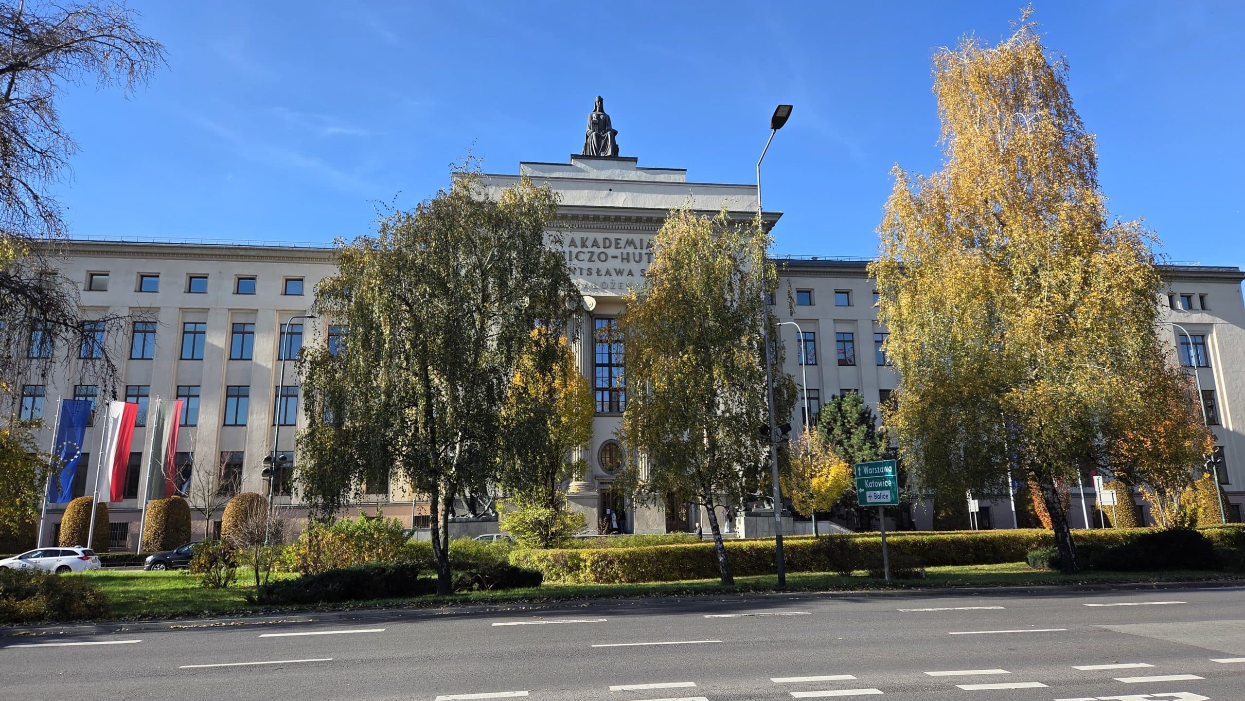 Ein großes akademisches Gebäude mit einer Statue auf dem Dach, polnische Fahnen auf der linken Seite und hohe Bäume mit Herbstlaub davor. Der Himmel ist klar und blau, und entlang der Straße sind Autos geparkt.