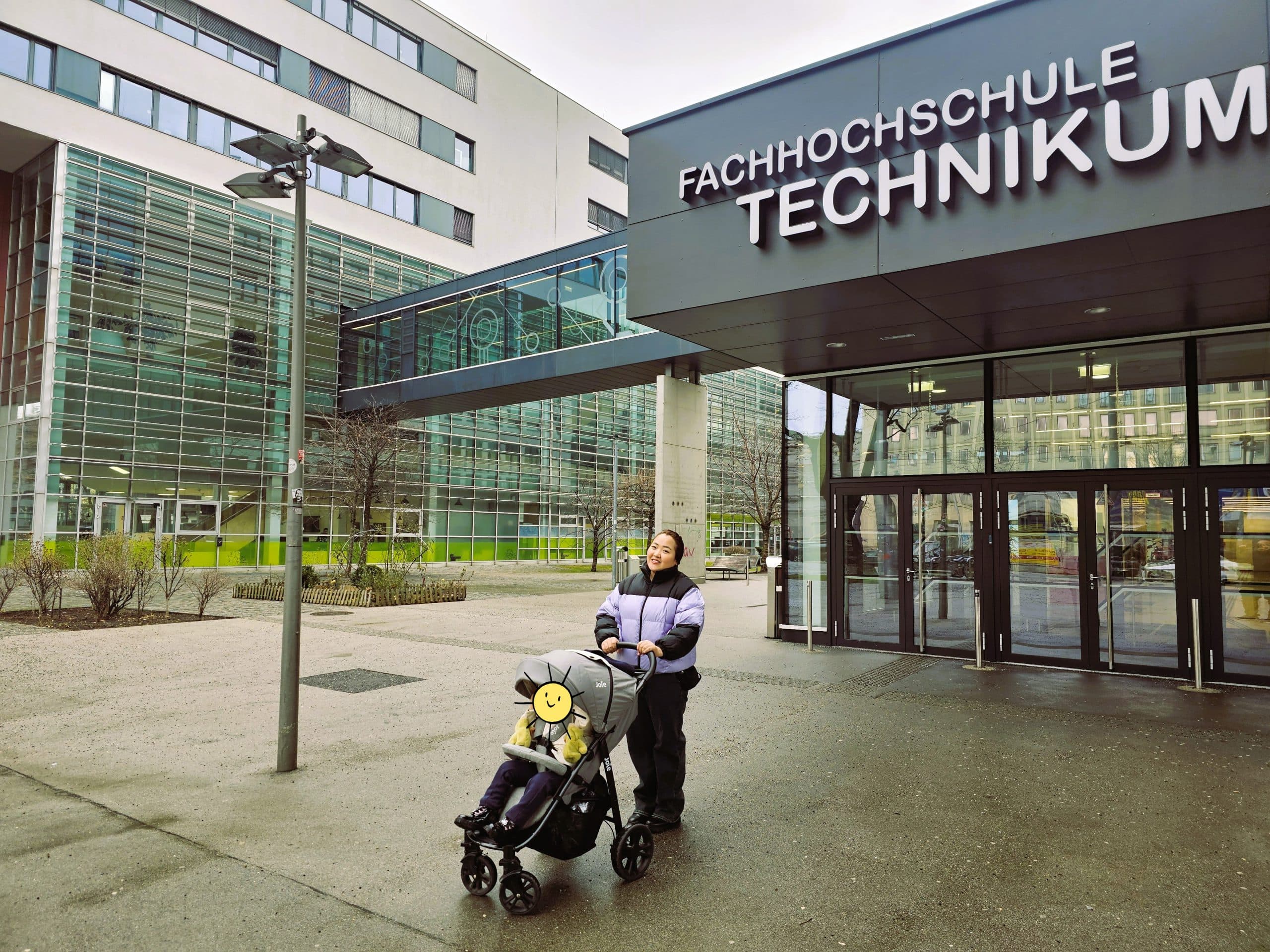 Eine Person steht vor dem Eingang der Fachhochschule Technikum und schiebt einen Kinderwagen mit einem Kind, das ein Smiley-Emoji im Gesicht hat. Im Hintergrund sind moderne Glasgebäude zu sehen.
