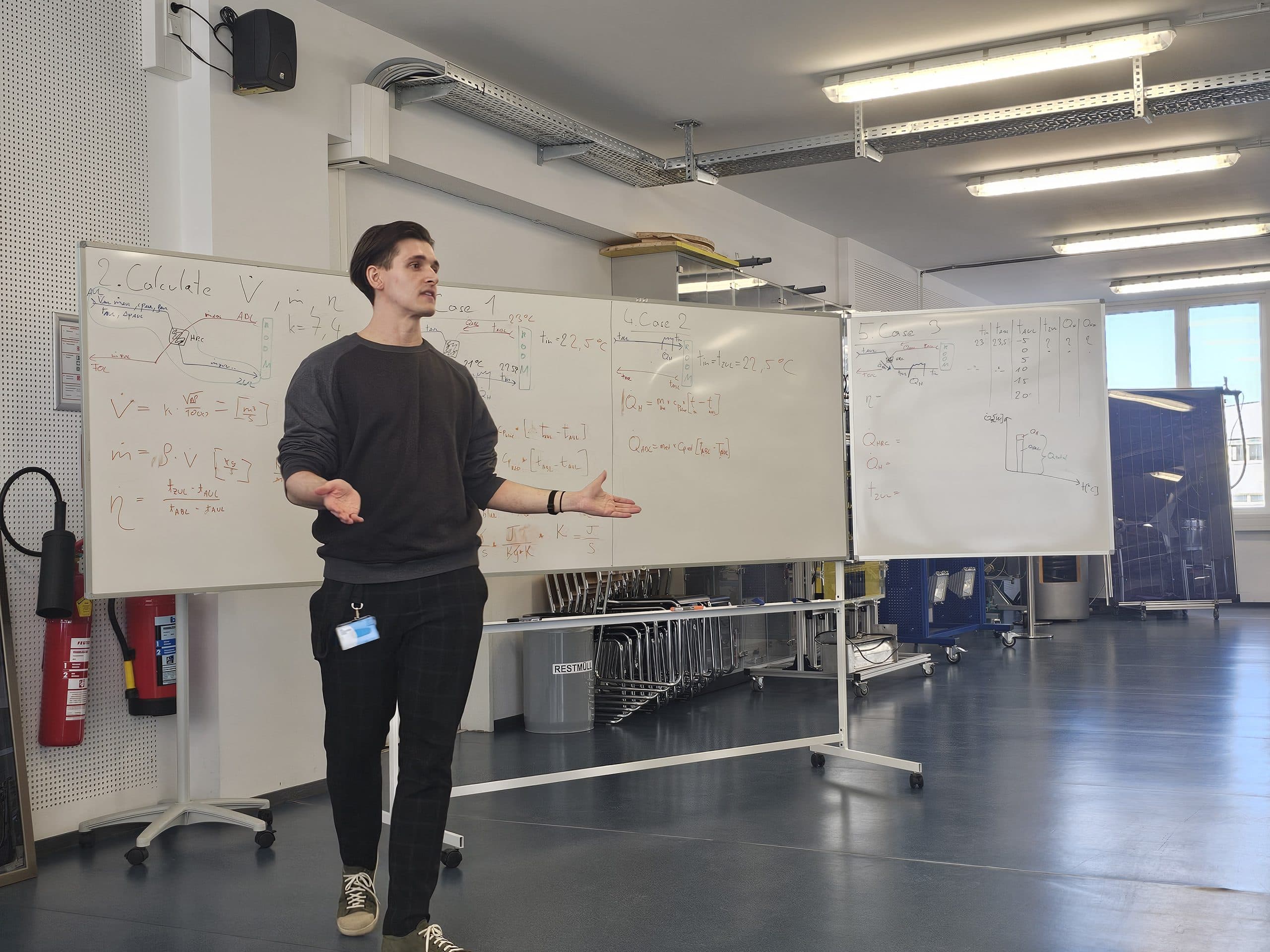 Eine Person steht in einem hellen Klassenzimmer mit blauem Fußboden, Tischen und Stühlen vor zwei Whiteboards mit mathematischen Gleichungen und gestikuliert mit beiden Händen.