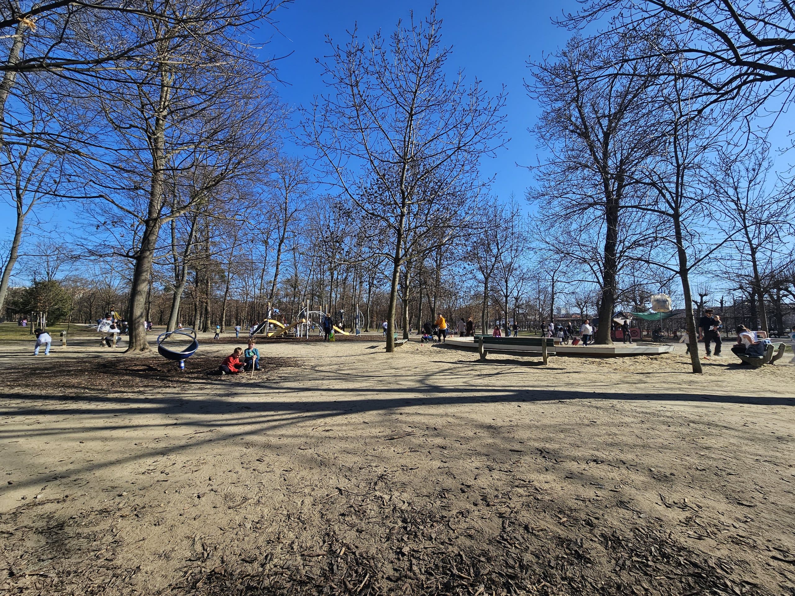 Menschen genießen einen sonnigen Tag in einem Park mit kahlen Bäumen, einige sitzen auf Bänken, Kinder spielen, andere entspannen sich in einer Hängematte. Der Himmel ist klar und blau, was auf einen frühen Frühling oder Spätherbst hindeutet.