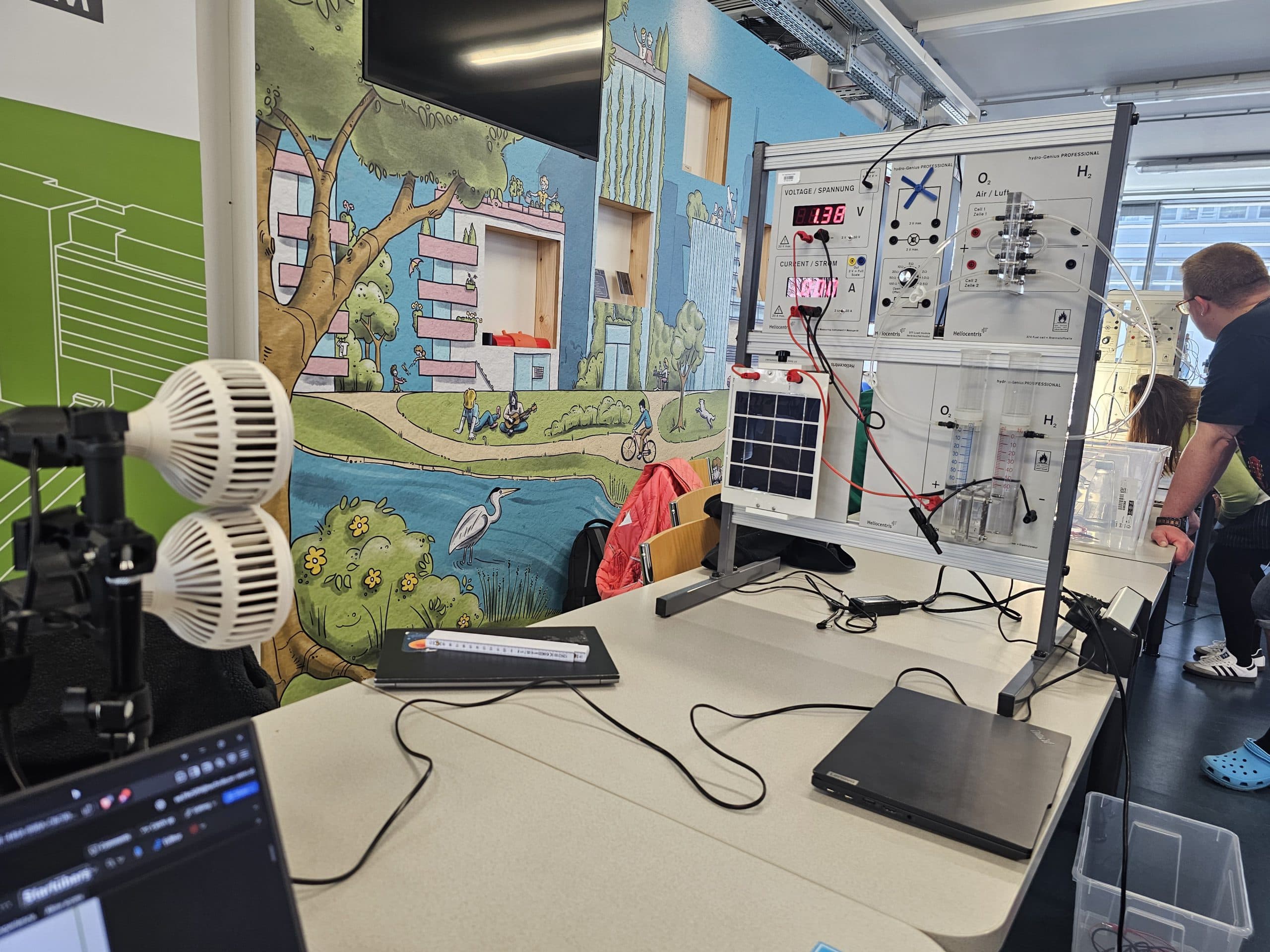 Ein wissenschaftlicher Versuchsaufbau mit elektronischen Geräten und Sensoren auf einem Tisch, einem Laptop und einer bunten Wandmalerei von Natur und Gebäuden an der Wand dahinter. Im Hintergrund arbeiten Menschen.