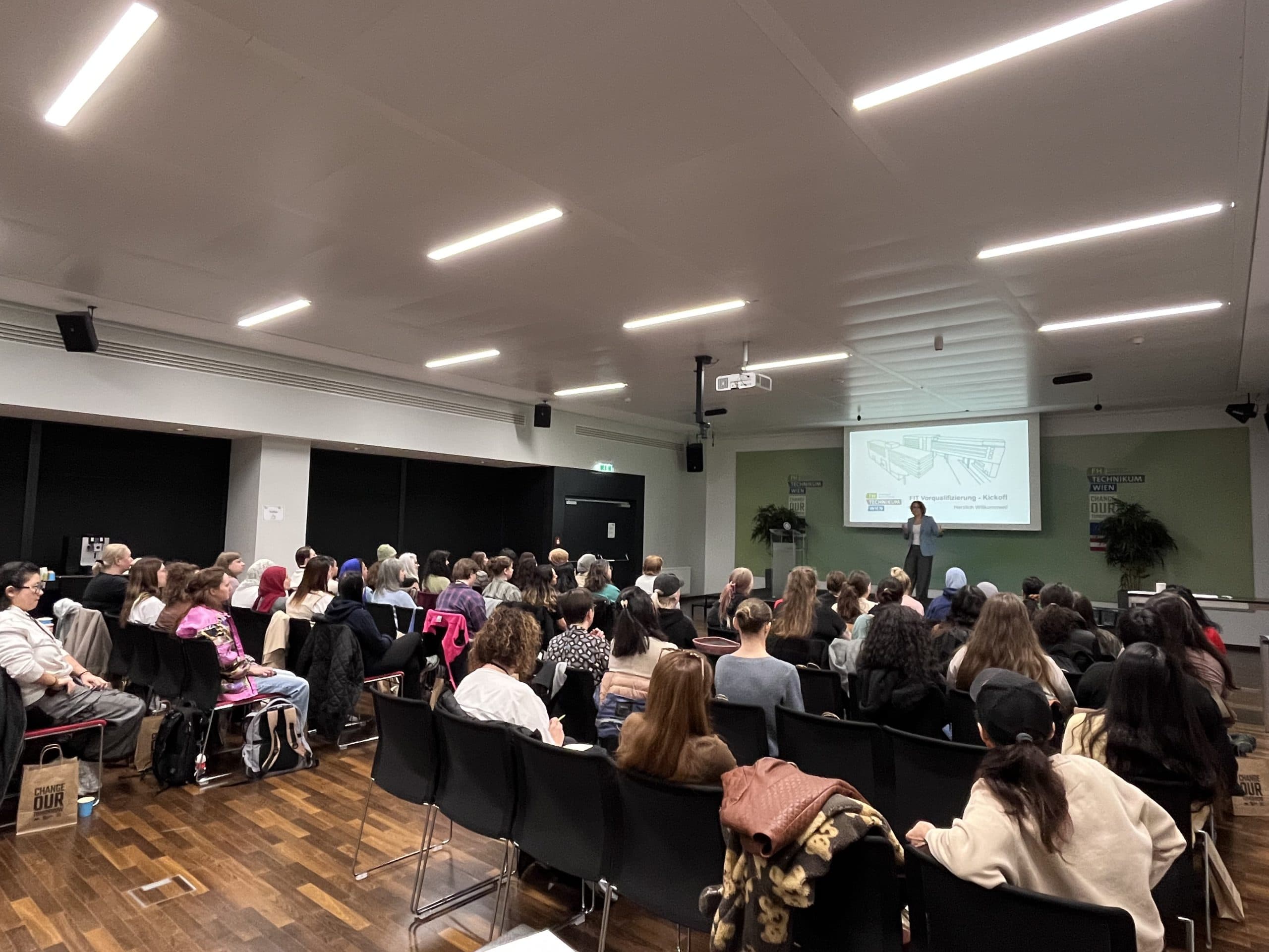 Eine große Gruppe von Personen sitzt in Reihen vor einem Moderator in einem modernen Konferenzraum und sieht sich ein projiziertes Dia auf der Leinwand an. Der Raum hat Holzböden und weiße Decken.