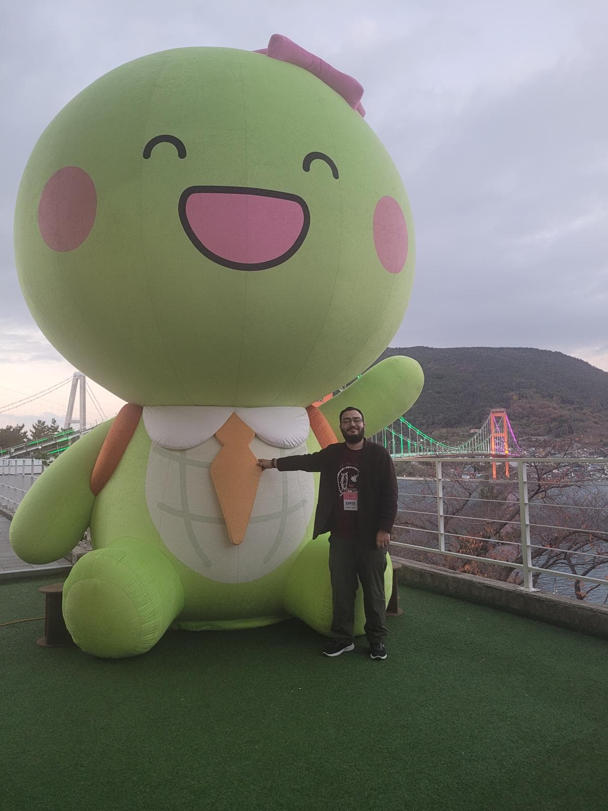 Eine Person steht lächelnd neben einer großen grünen Zeichentrickfigur mit Krawatte und Hut, im Freien mit einer bunten Brücke und Hügeln im Hintergrund.