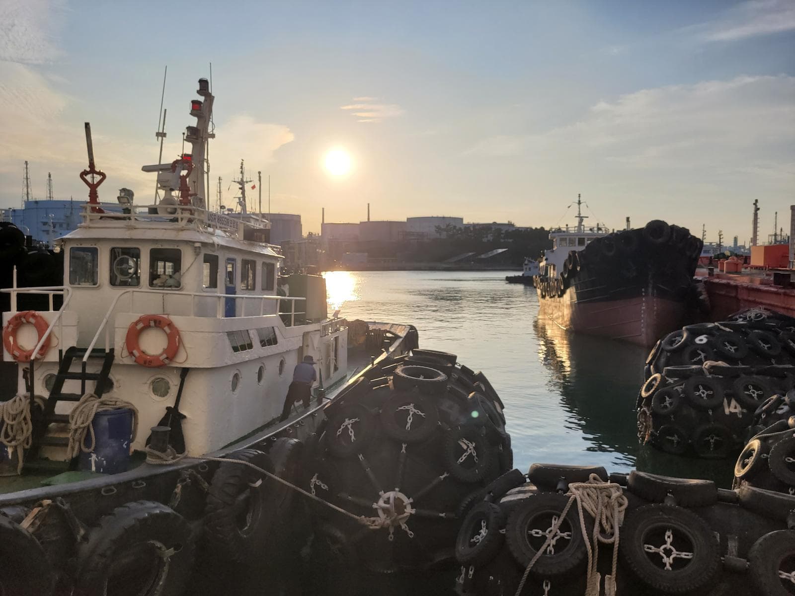 An einem Hafen angedockte Boote bei Sonnenuntergang, wobei sich das Sonnenlicht auf dem Wasser spiegelt und große schwarze Fender an den Booten befestigt sind. Im Hintergrund sind Gebäude und Kräne bei teilweise bewölktem Himmel zu sehen.