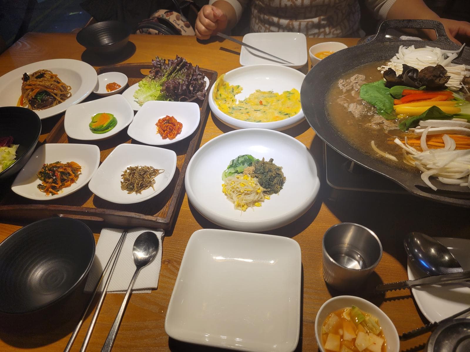 Ein Tisch mit verschiedenen koreanischen Gerichten, darunter Beilagen, eine große Pfanne mit Gemüse und Brühe, kleine Teller mit Kimchi, Grünzeug und Gewürzen, um den herum Utensilien und Servietten angeordnet sind.