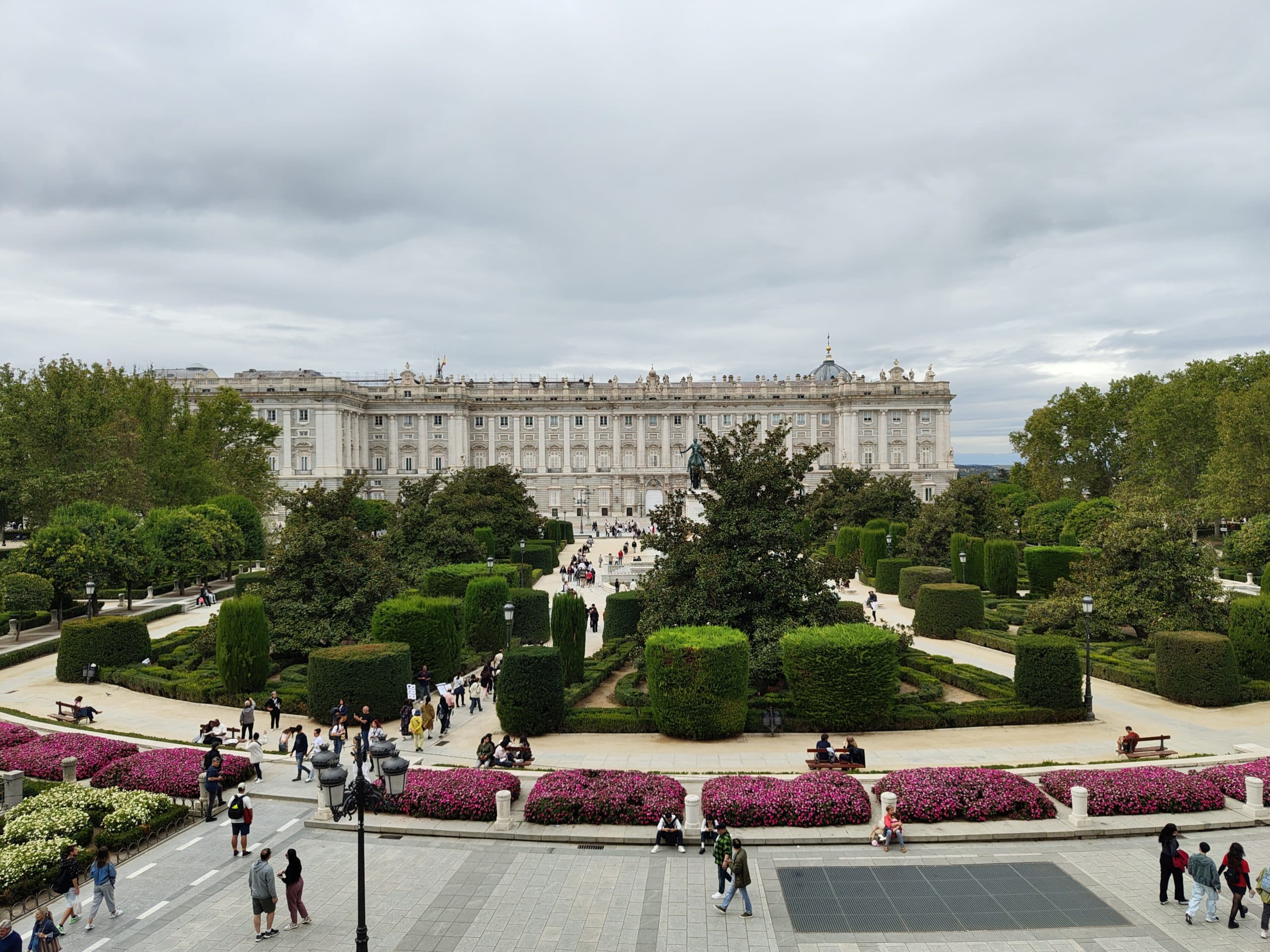 Das Bild zeigt den Königspalast von Madrid hinter üppigen formalen Gärten mit gestutzten Hecken, blühenden Pflanzen und Menschen, die unter einem wolkenverhangenen Himmel über Wege spazieren.