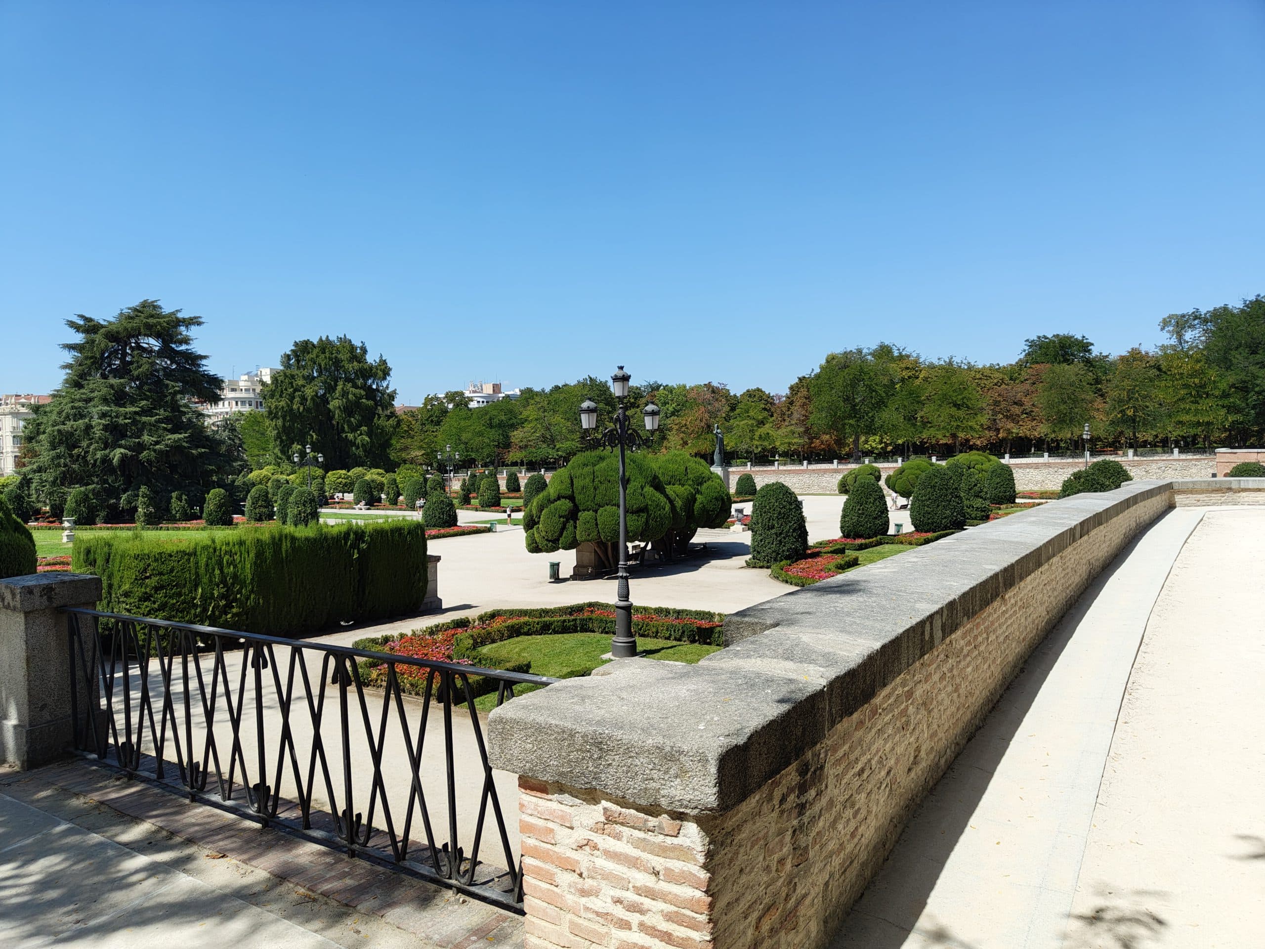 Ein sonniger Tag in einem formalen Park mit ordentlich getrimmten Büschen, Bäumen, Blumenbeeten und einem schwarzen Eisenzaun entlang einer Steinmauer. Im Hintergrund sind der strahlend blaue Himmel und die Wege zu sehen.