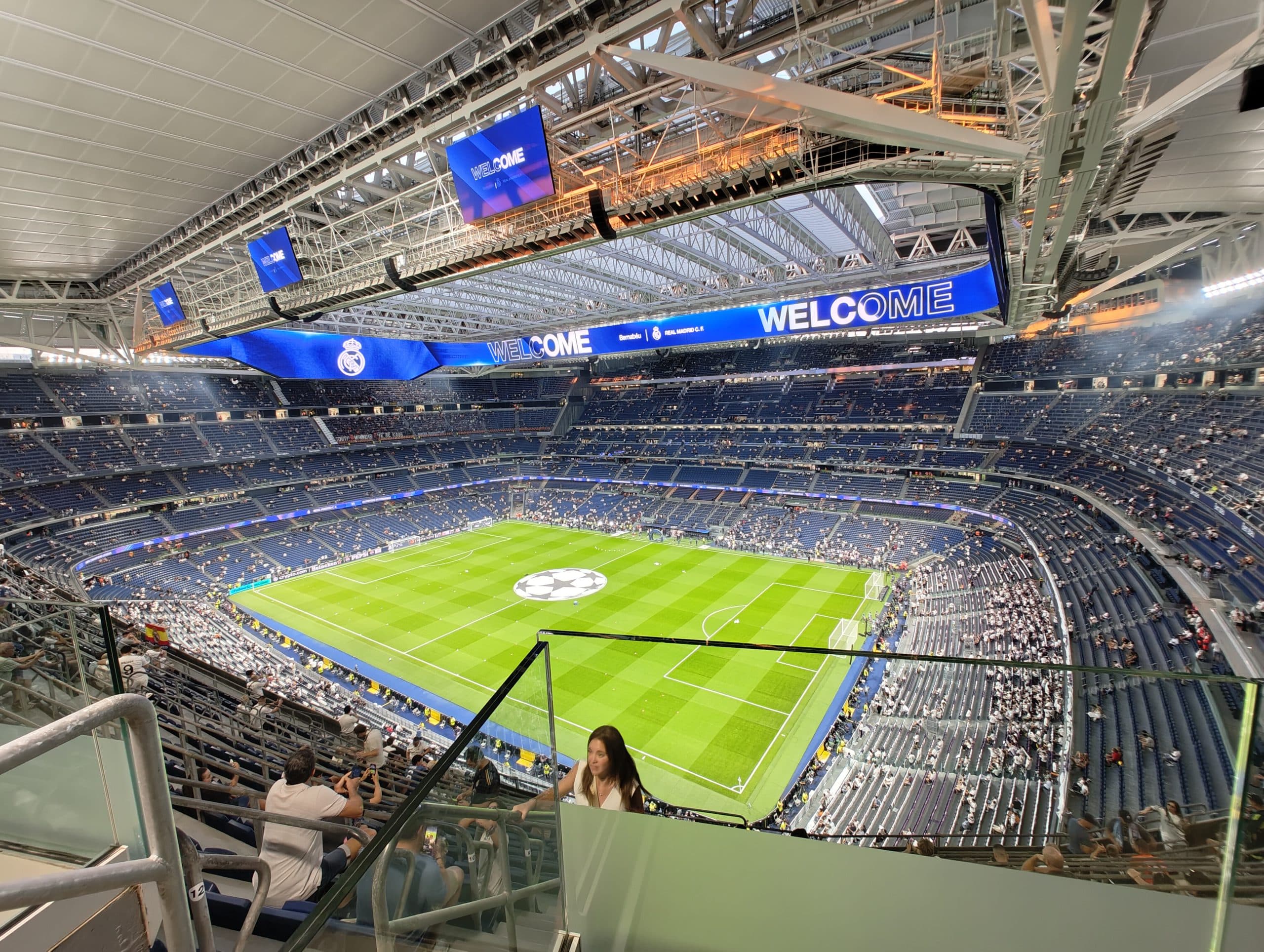 Weitwinkelansicht eines großen, modernen Stadions mit einem einziehbaren Dach, das mit Zuschauern gefüllt ist. Das Spielfeld ist für ein Fußballspiel vorbereitet, und auf den großen Bildschirmen wird "WELCOME" angezeigt.