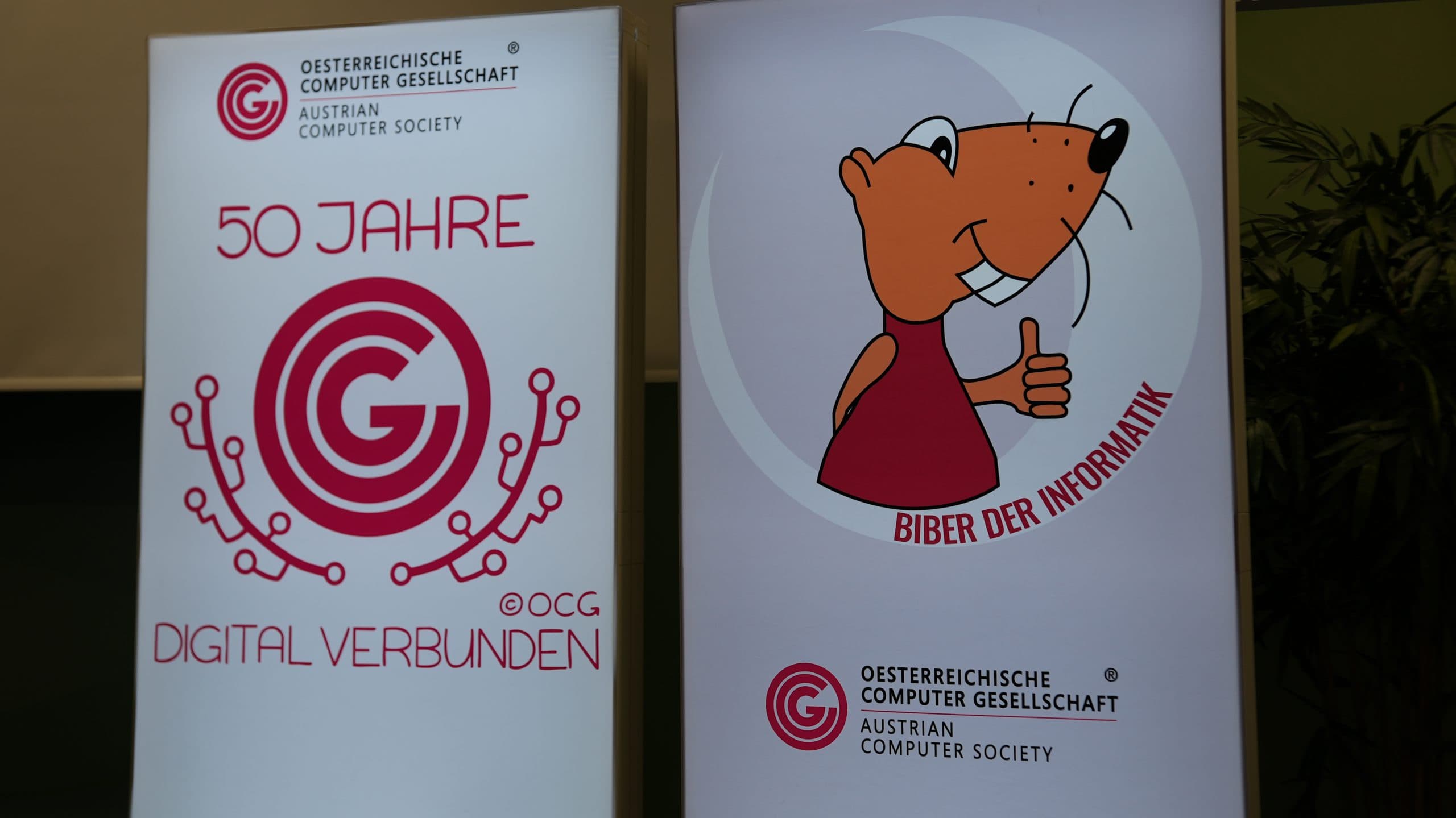 Zwei Transparente für die Österreichische Computergesellschaft. Das linke Banner trägt die Aufschrift '50 Jahre Digital Verbunden' mit einem Logo mit technischem Bezug. Das rechte Banner zeigt einen Cartoon-Biber, der den Daumen nach oben streckt, und den Text 'Biber der Informatik'.