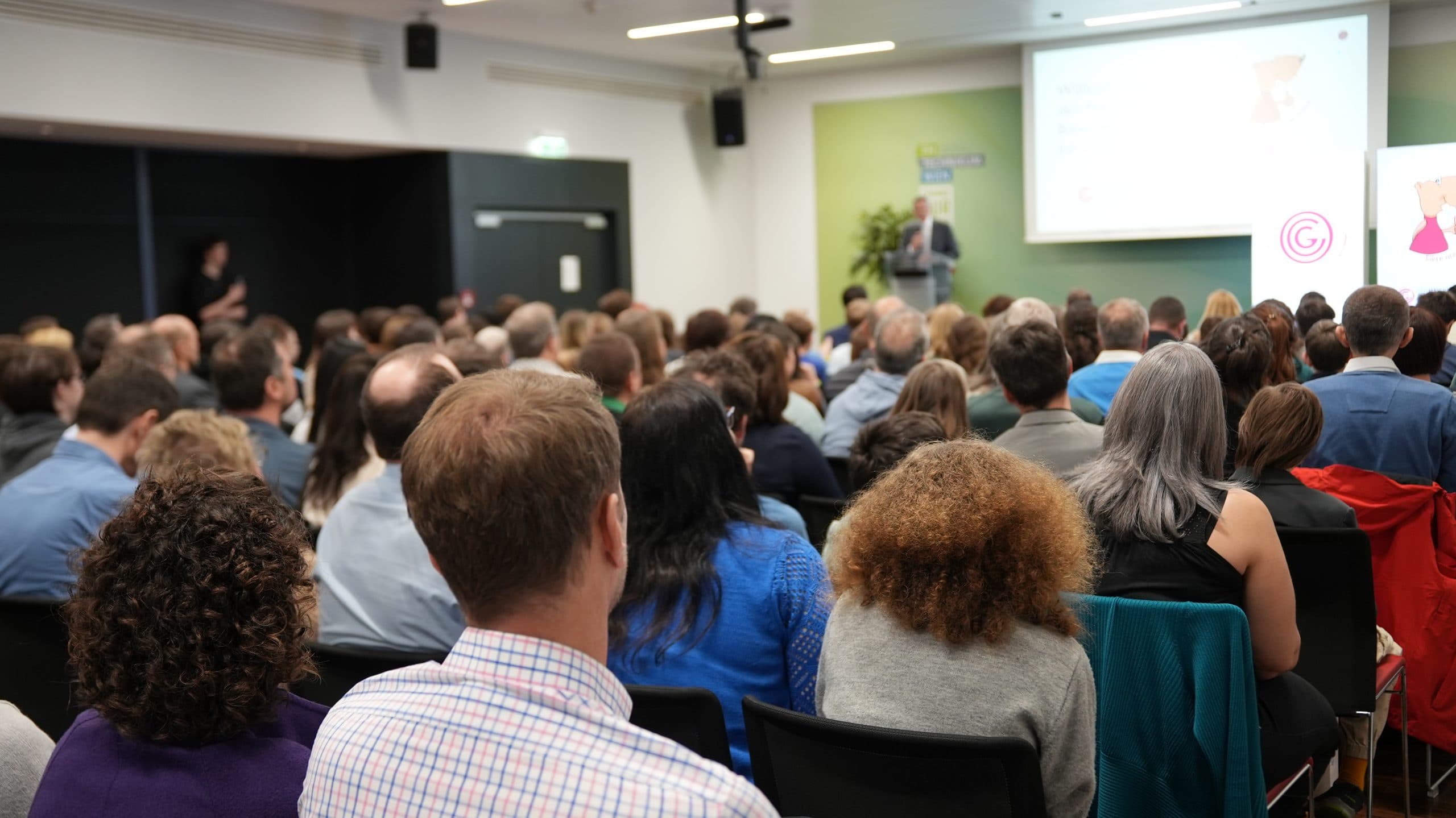 Ein großes Publikum sitzt in Reihen vor einem Redner auf einem Podium in einem modernen Konferenzraum und hört einer Präsentation zu, die auf eine Leinwand projiziert wird.