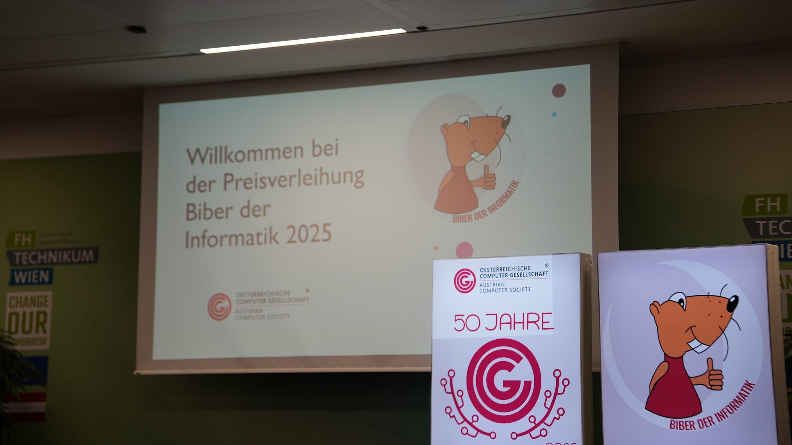 Eine Projektionsfläche zeigt Willkommen bei der Preisverleihung Biber der Informatik 2025 mit einem Cartoon-Biber-Logo. Davor steht ein Aufsteller mit der Aufschrift 50 Jahre und dem gleichen Biber und Logo.