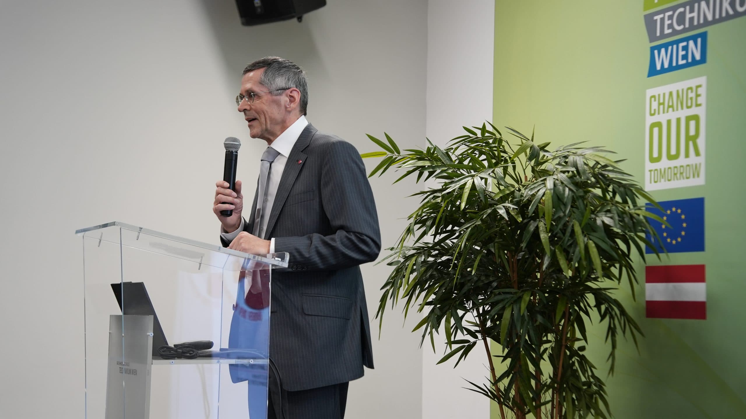 Ein Mann im Anzug spricht an einem durchsichtigen Podium mit einem Mikrofon. Hinter ihm befindet sich eine grüne Wand mit Pflanzen und Schildern mit den Aufschriften Technik, Wien und Change Our Tomorrow sowie EU- und Österreich-Flaggen.