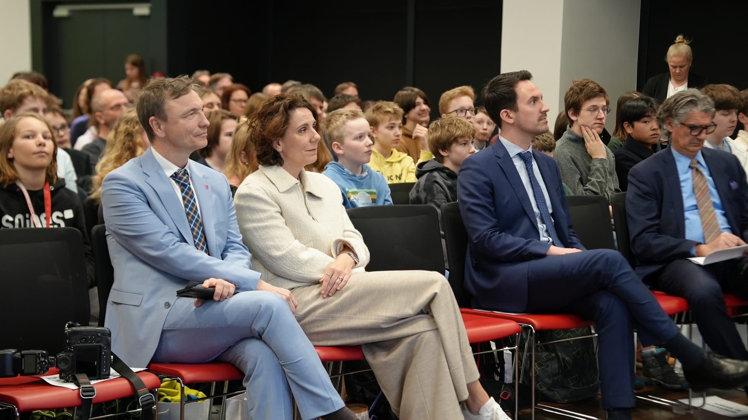 Eine Gruppe formal gekleideter Erwachsener sitzt in der ersten Reihe eines Hörsaals, während hinter ihnen ein großes Publikum, meist Kinder und Jugendliche, aufmerksam eine Veranstaltung verfolgt.