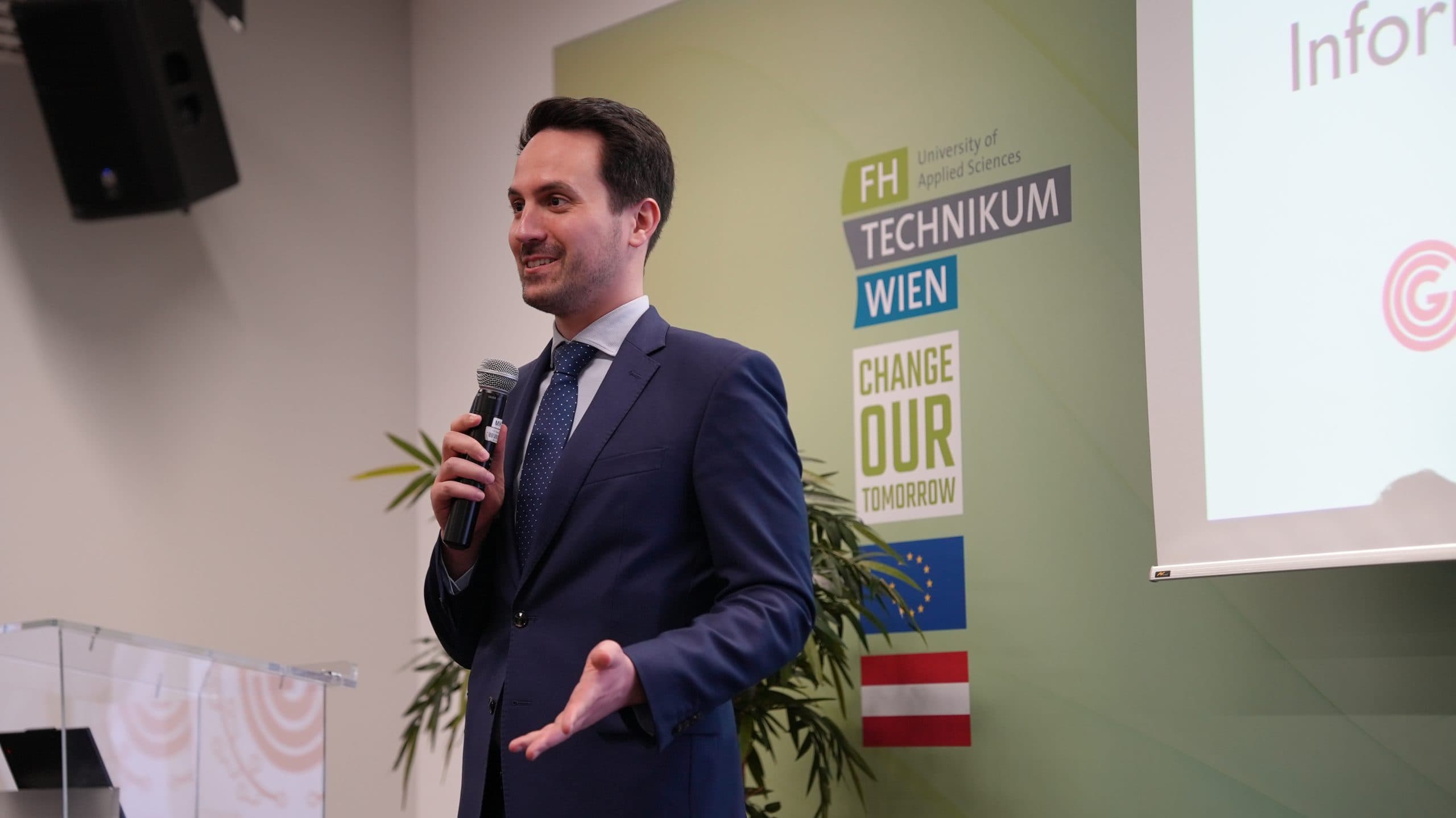 Ein Mann in dunklem Anzug und Krawatte spricht von einem Podium aus in ein Mikrofon. Hinter ihm ist eine grüne Wand mit dem Branding der FH Technikum Wien, der österreichischen Flagge und dem Slogan Change Our Tomorrow zu sehen. Auf der rechten Seite ist teilweise ein Bildschirm zu sehen.