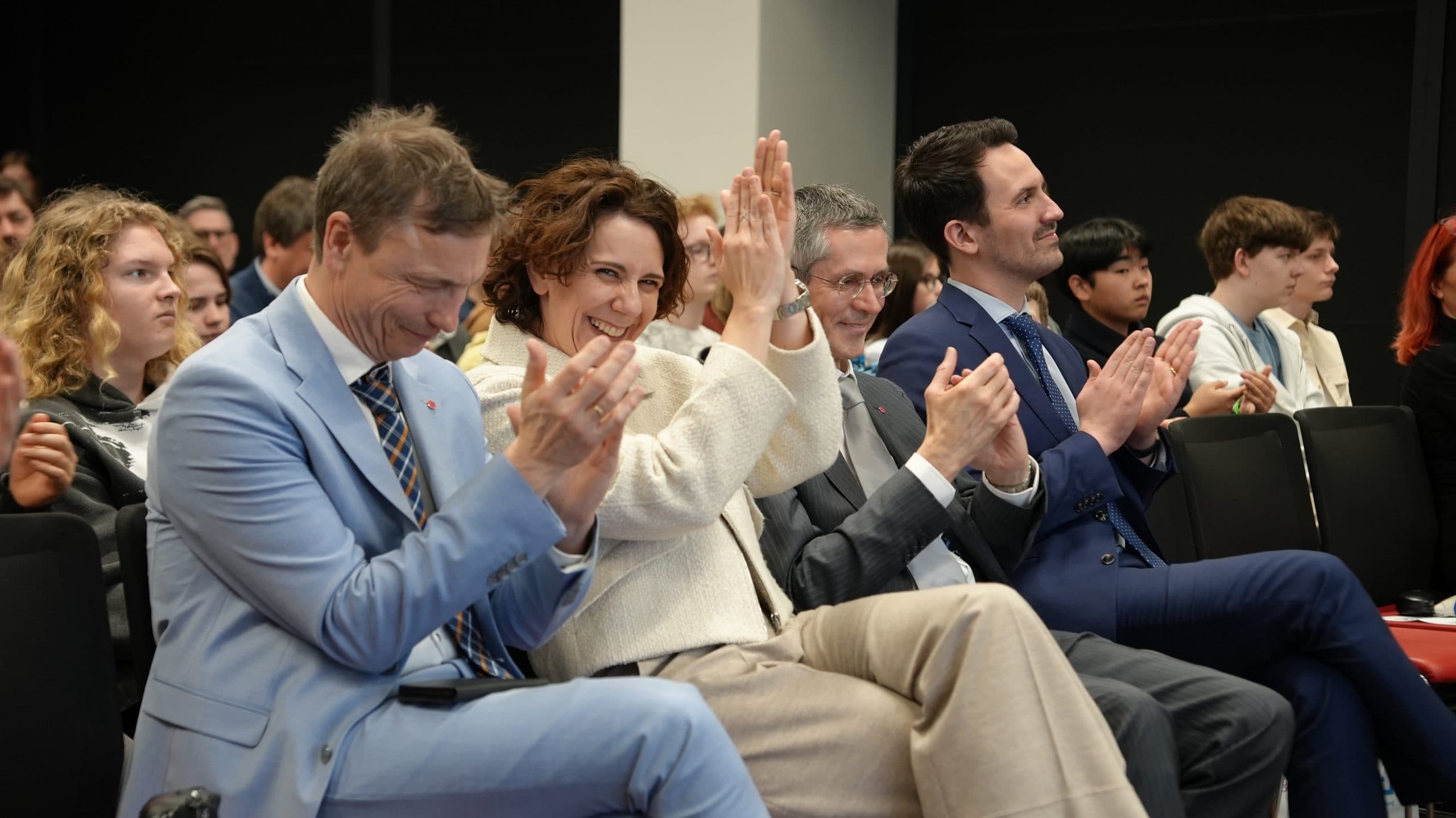 Eine Gruppe formell gekleideter Personen sitzt in Reihen und klatscht enthusiastisch, lächelt und zeigt ihre Wertschätzung während einer Veranstaltung oder Präsentation. Das Publikum ist vielfältig und engagiert.