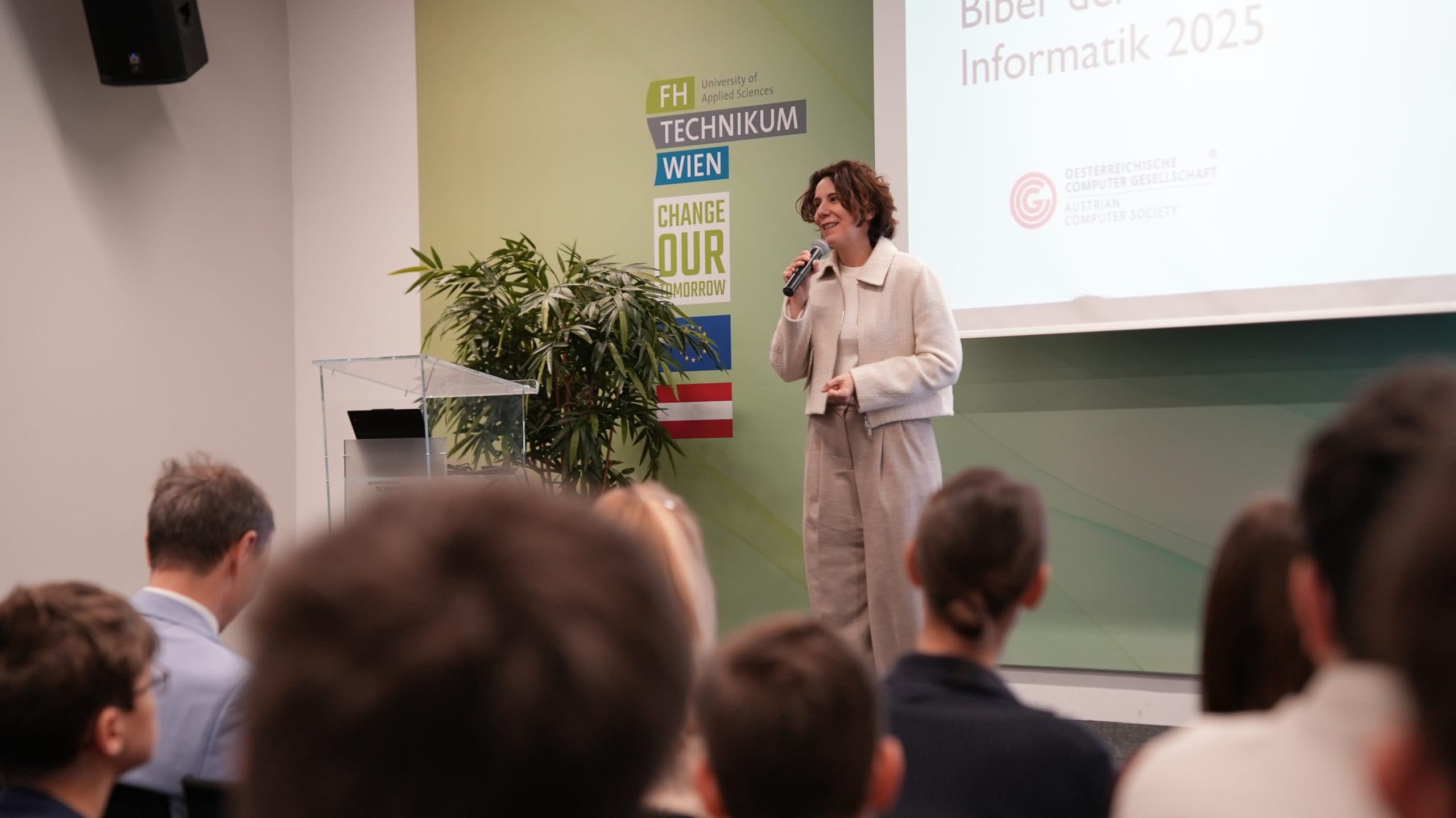 Eine Frau spricht auf der Bühne vor einem Publikum in ein Mikrofon, während hinter ihr eine Präsentation projiziert wird und an der Wand eine Pflanze und Universitätsbanner zu sehen sind.