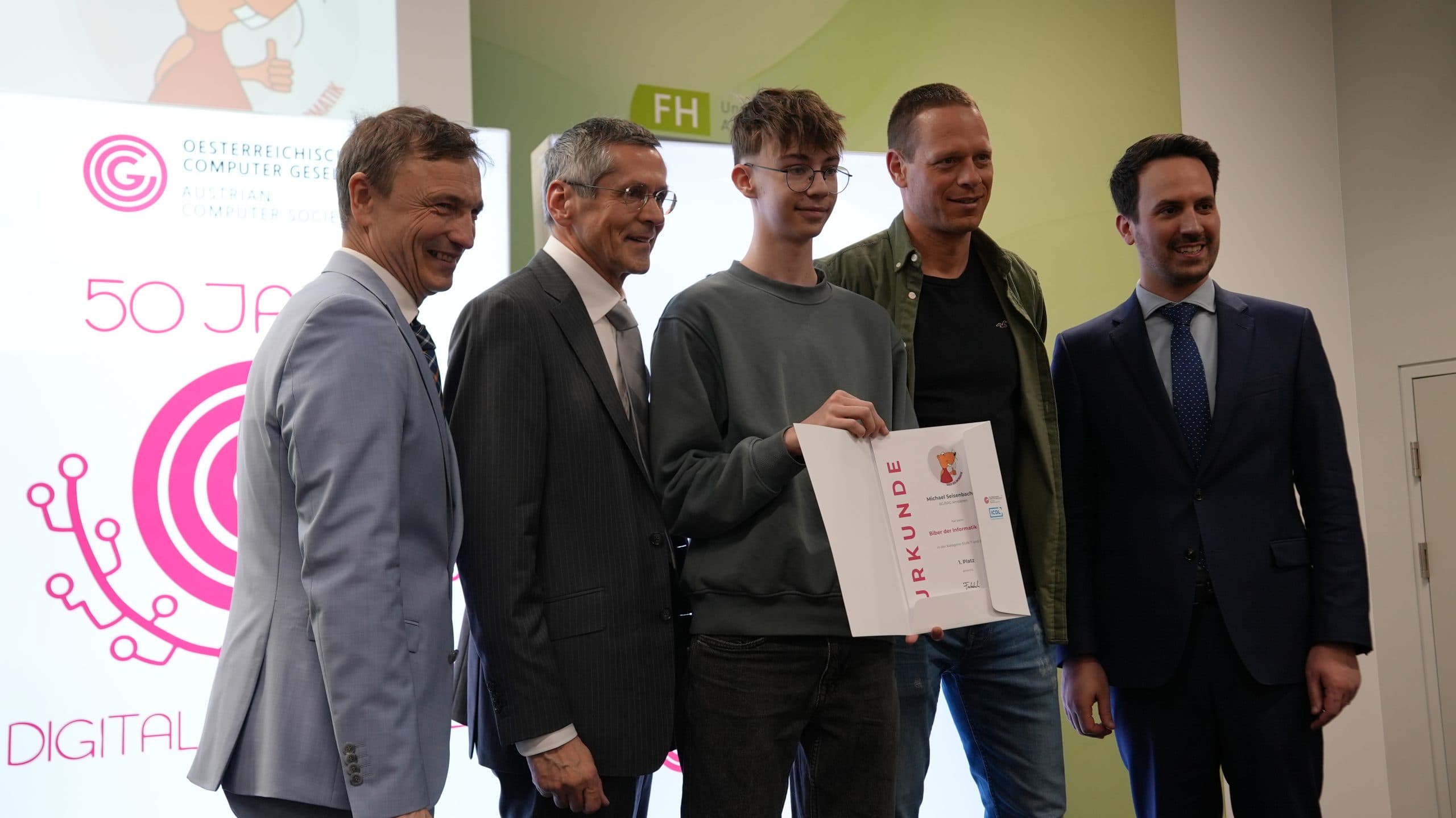 Ein junger Mensch mit einer Urkunde steht in der Mitte von vier Erwachsenen in Anzügen und Freizeitkleidung, die für ein Foto bei einer Preisverleihung mit Veranstaltungsbannern im Hintergrund posieren und lächeln.