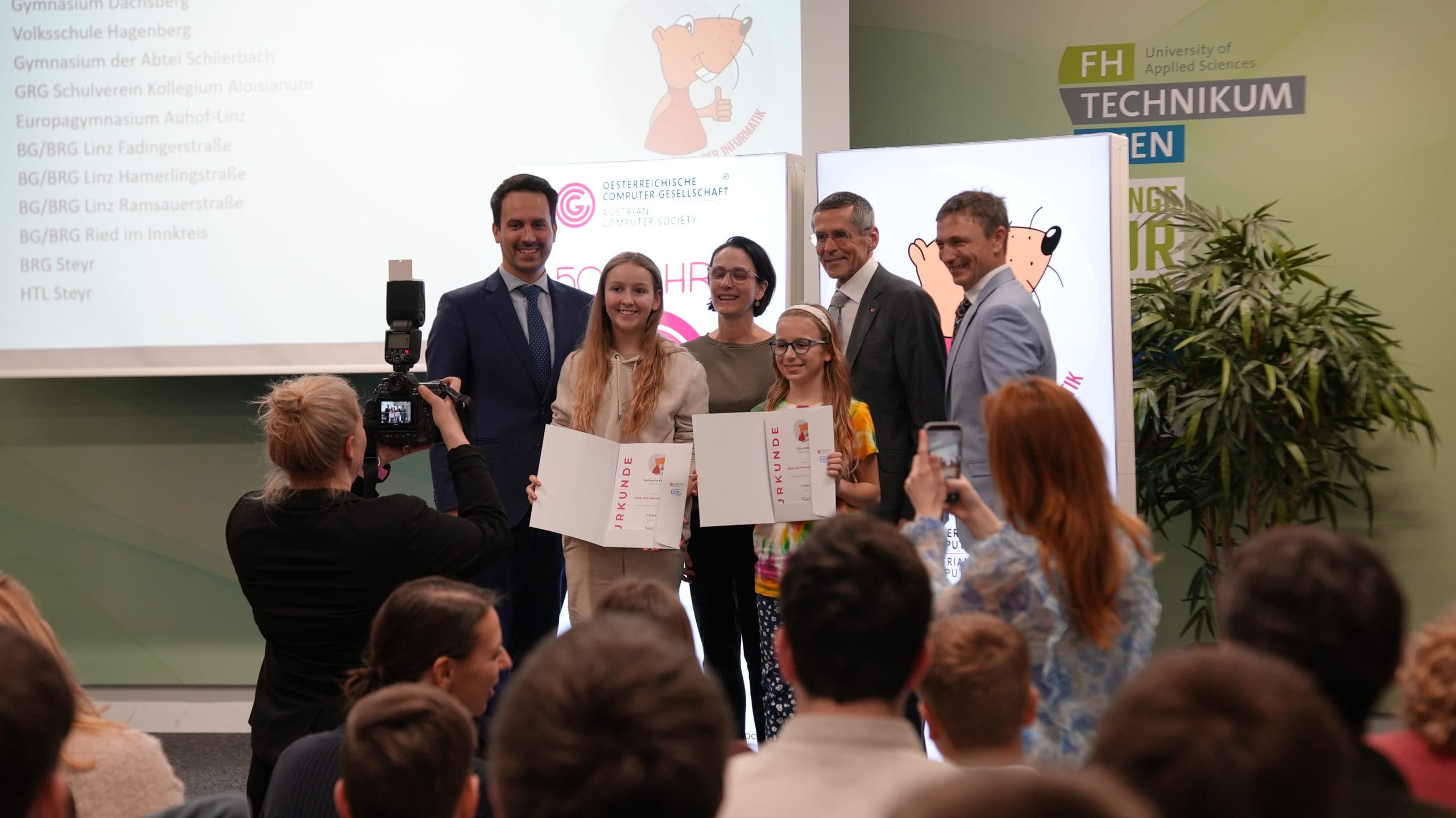 Eine Gruppe von Studenten und Erwachsenen posiert auf der Bühne mit Urkunden vor einem Publikum, während jemand ein Foto von ihnen macht. Im Hintergrund wird ein Zeichentrickbär auf die Leinwand projiziert, auf der die Logos der Universität und der Veranstaltung zu sehen sind.