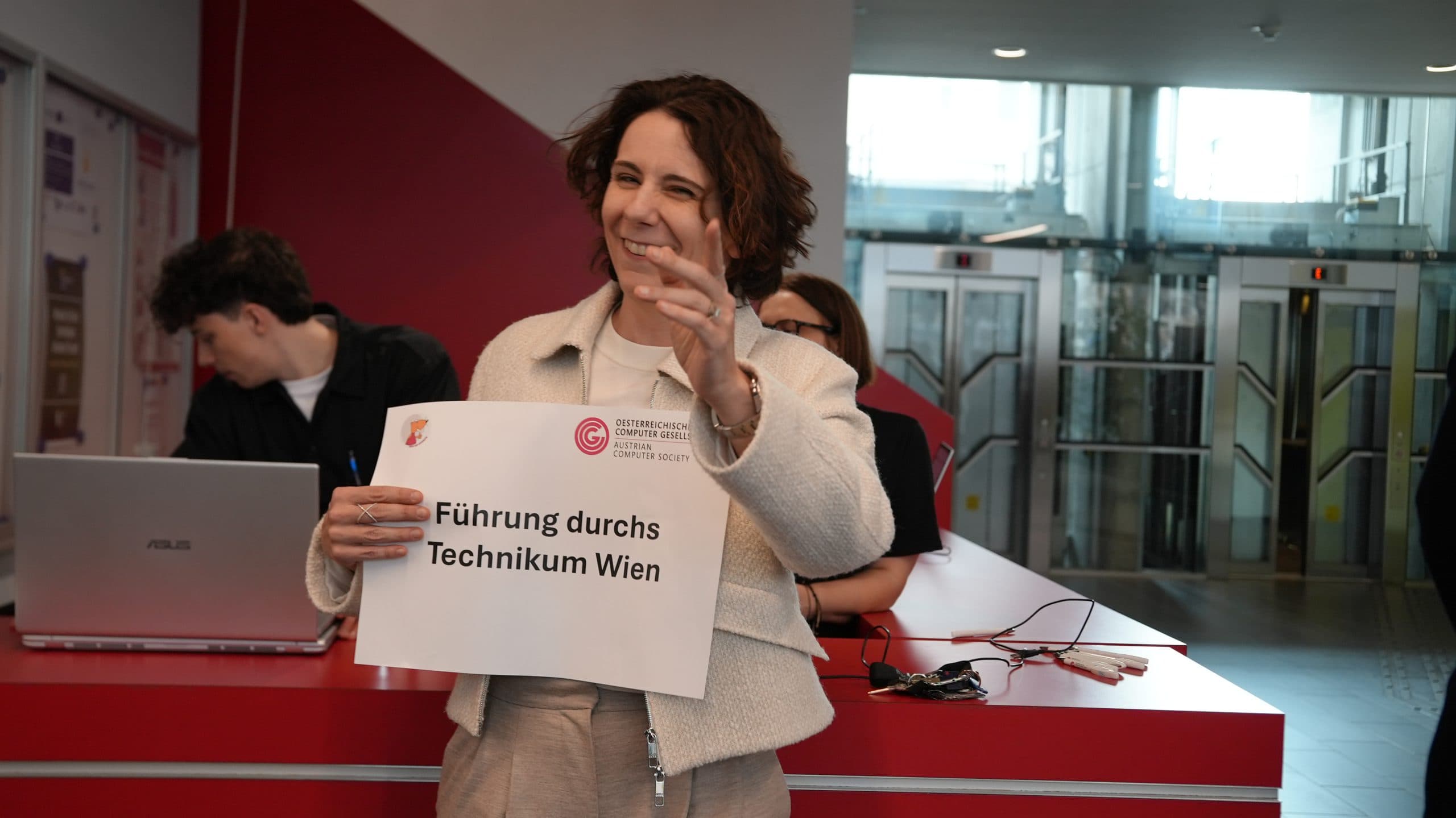 Eine Frau steht lächelnd in einem Raum und hält ein Schild mit der Aufschrift Führung durchs Technikum Wien. Hinter ihr arbeitet eine Person an einem Laptop an einem roten Schreibtisch, und im Hintergrund sind Glastüren und Aufzüge zu sehen.