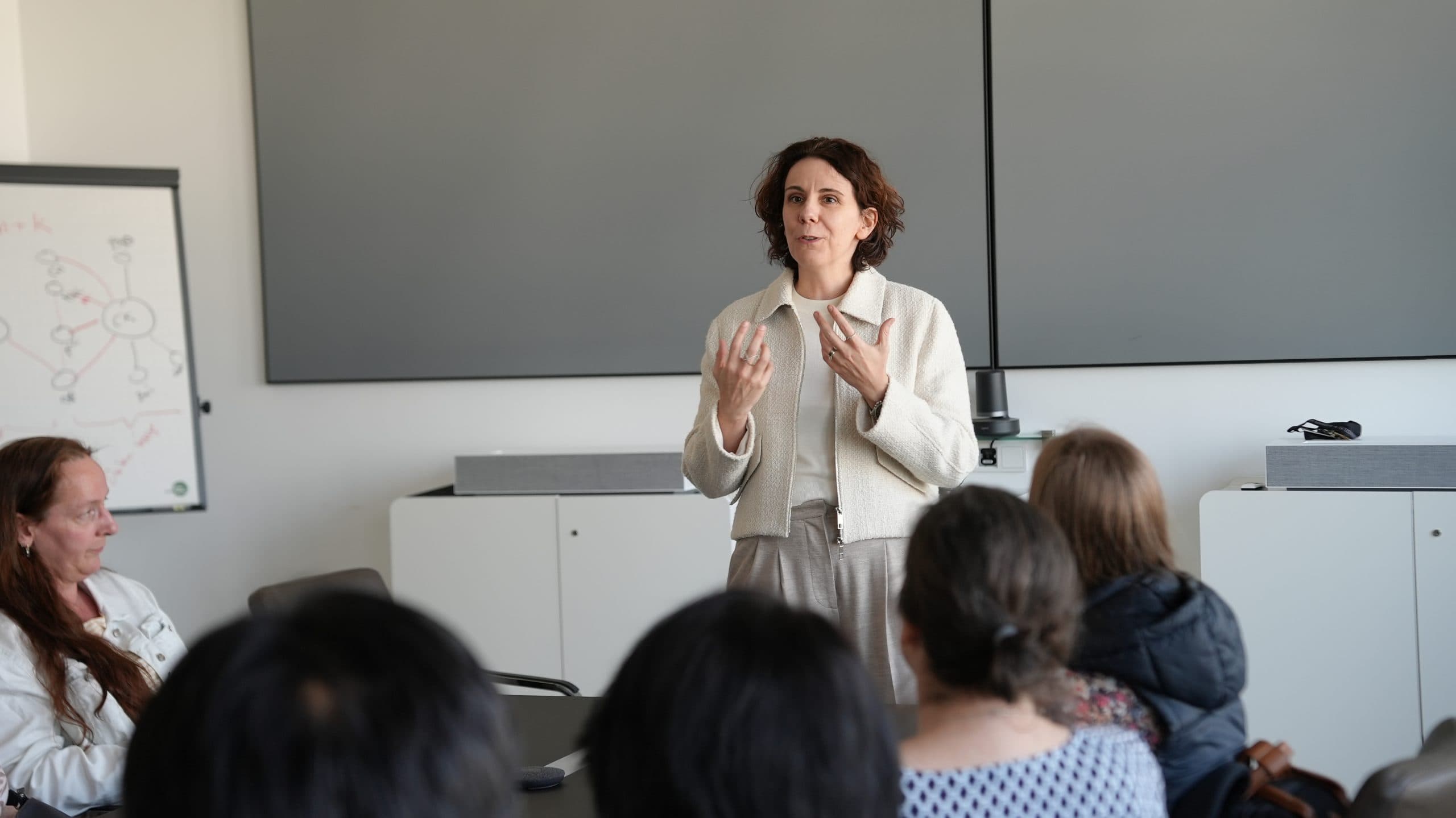 Eine Frau steht und spricht zu einer sitzenden Gruppe in einem modernen Konferenzraum, wobei sie mit den Händen gestikuliert. Mehrere Personen stehen ihr gegenüber und hören ihr aufmerksam zu. Im Hintergrund sind ein Whiteboard und große Bildschirme zu sehen.