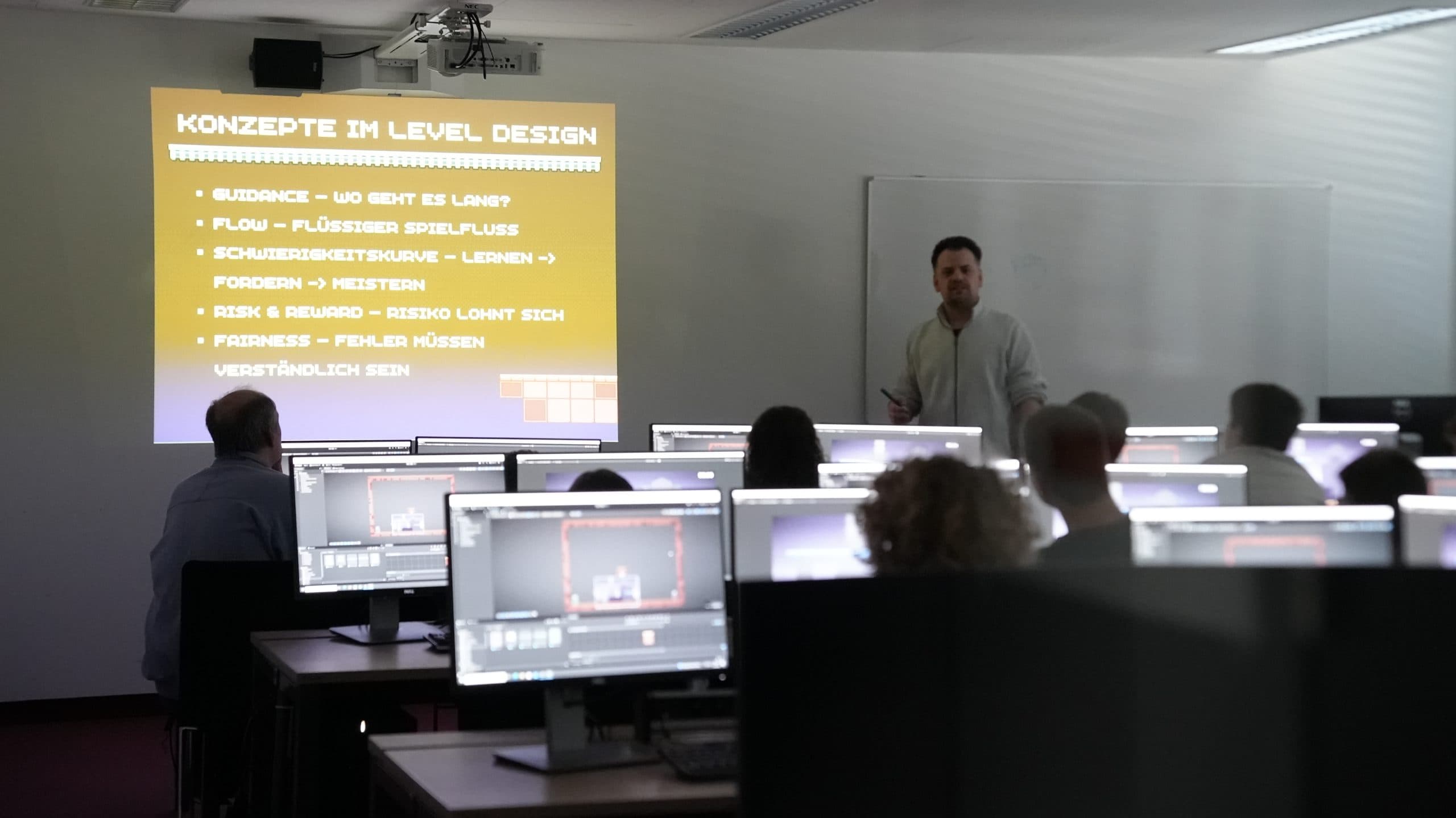 Ein Mann hält einen Vortrag über Konzepte im Level Design vor einem Klassenzimmer mit Schülern, die an Computerstationen arbeiten, wobei eine Folie mit den wichtigsten Begriffen auf Deutsch an die Wand projiziert wird.
