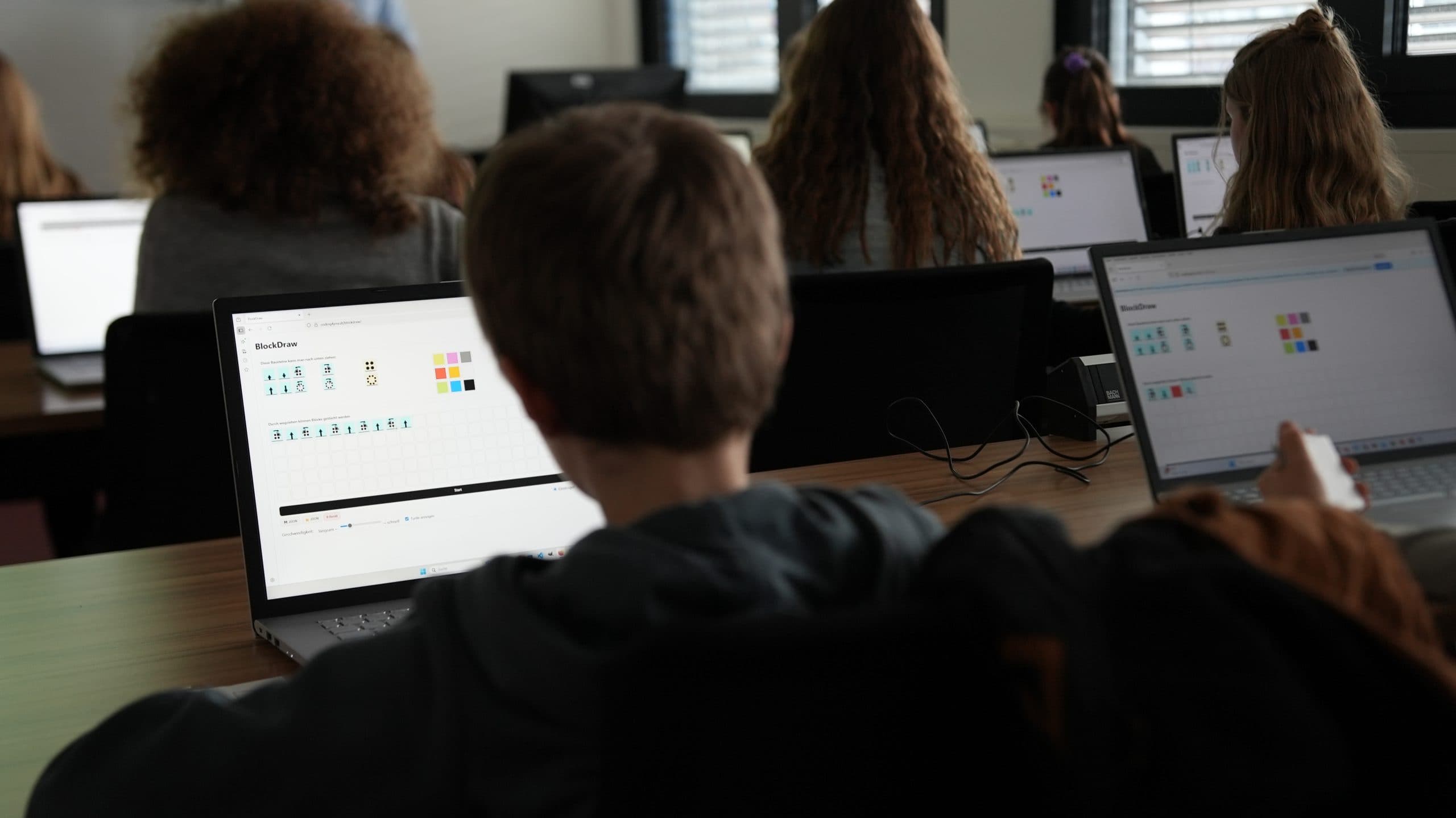 Schüler sitzen an Tischen in einem Klassenzimmer und arbeiten an Laptops. Das Hauptaugenmerk liegt auf einem Jungen, der eine Lernsoftware mit Symbolen und farbigen Formen auf seinem Bildschirm verwendet. Im Hintergrund sind andere Schüler zu sehen.