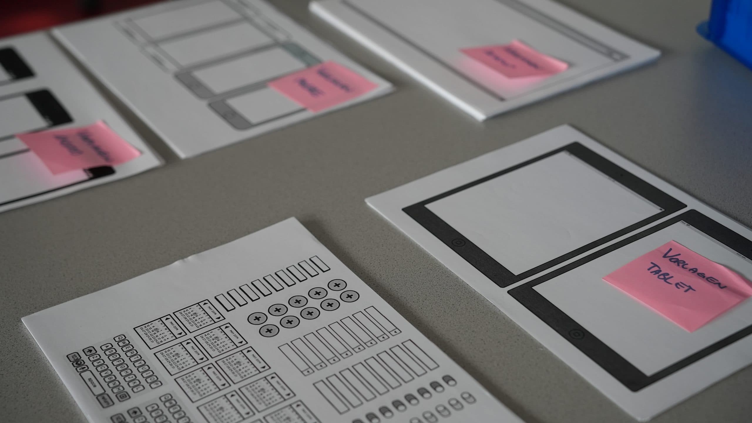 Auf einem Tisch liegen Papierbögen mit Wireframe-Skizzen von Benutzeroberflächen für mobile Anwendungen und rosa Haftnotizen, die einen Design- oder Planungsprozess für ein digitales Projekt andeuten.