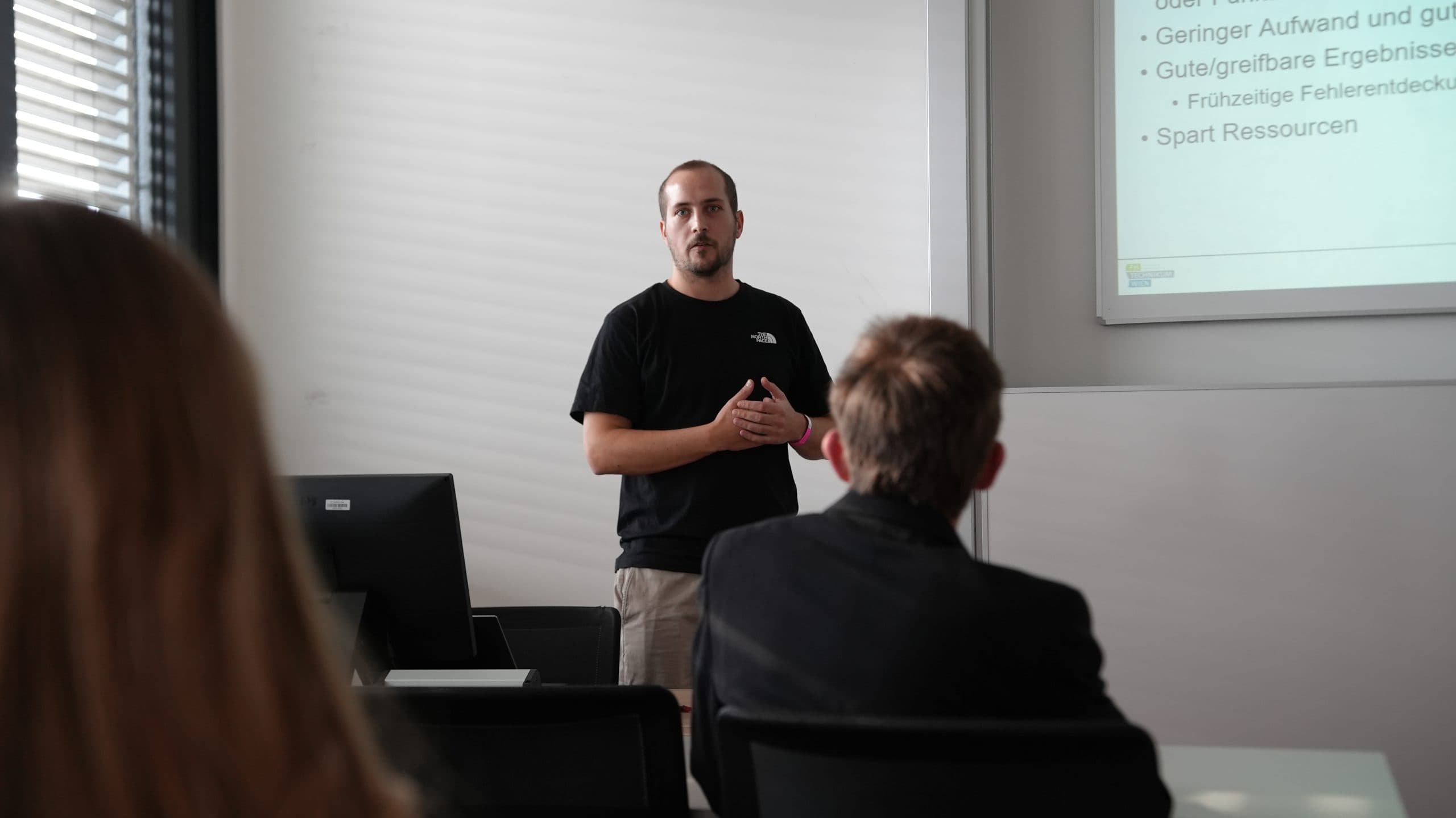 Ein Mann in einem schwarzen T-Shirt hält vor zwei sitzenden Personen in einem Klassenzimmer einen Vortrag, während an der Wand hinter ihm eine Folie mit deutschem Text projiziert wird.