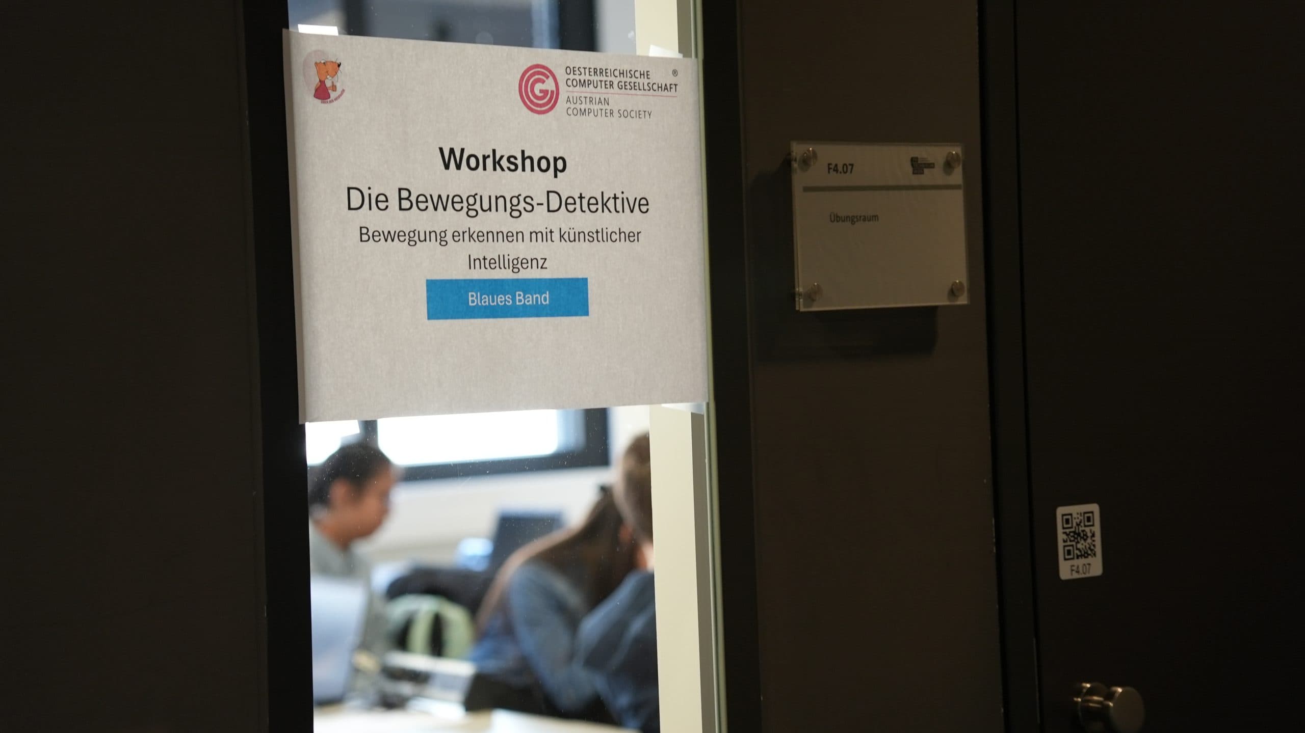 Auf einem Schild an einer Tür steht 'Workshop Die Bewegungs-Detektive: Bewegung erkennen mit künstlicher Intelligenz', im Hintergrund sitzen und arbeiten Menschen in einem Raum.