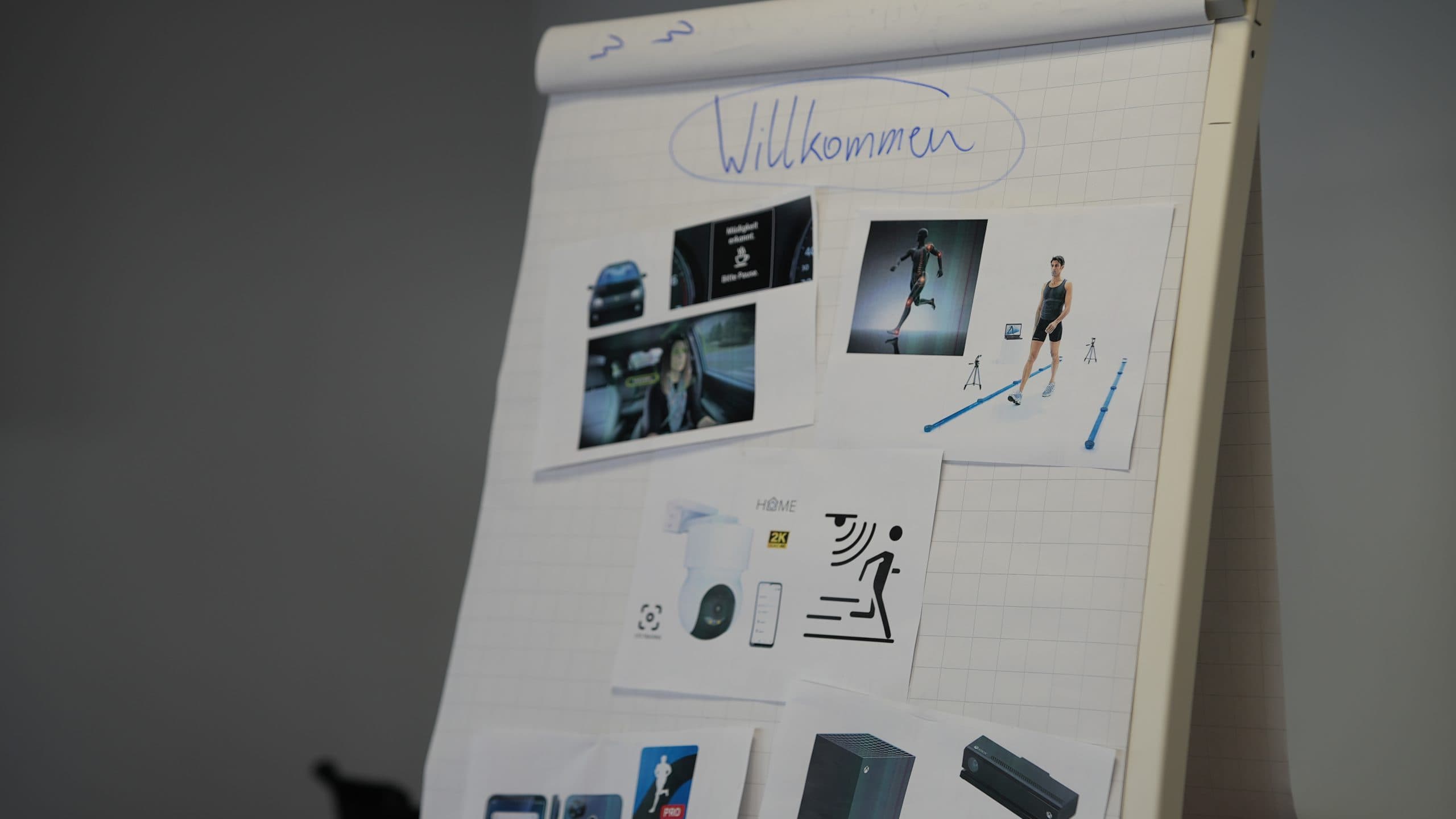 Ein Flipchart mit der Aufschrift 'Willkommen' am oberen Rand, auf dem mehrere gedruckte Bilder zu sehen sind, darunter Autos, eine Frau mit einem Headset, ein Läufer, Smart-Home-Geräte und elektronische Produkte.