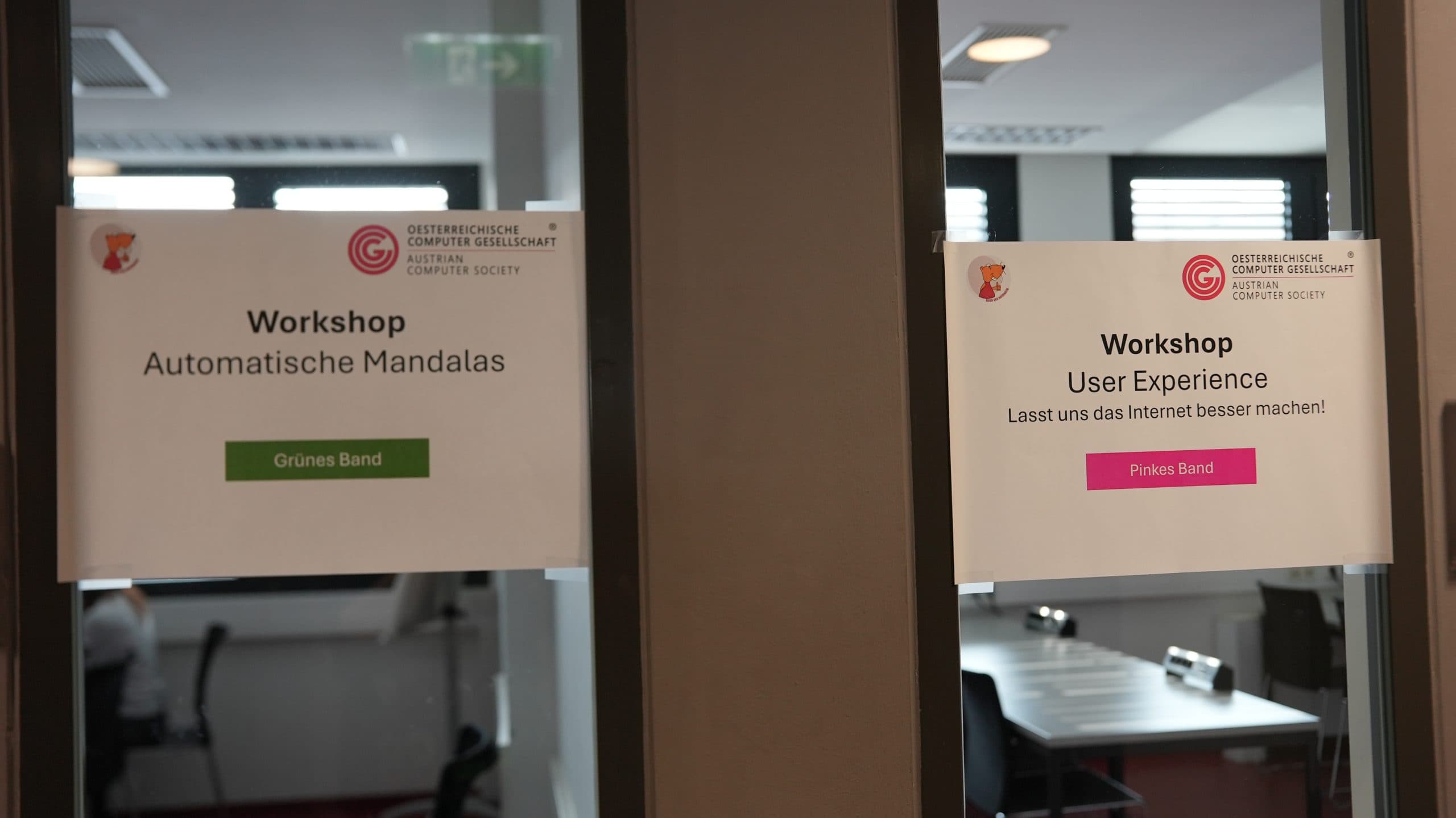Zwei Werkstattschilder in deutscher Sprache sind an Glastüren angebracht. Das linke Schild ist für Automatische Mandalas mit einem grünen Band, das rechte für User Experience mit einem rosa Band. Beide sind von der Computer Society. Im Inneren sind Schreibtische zu sehen.