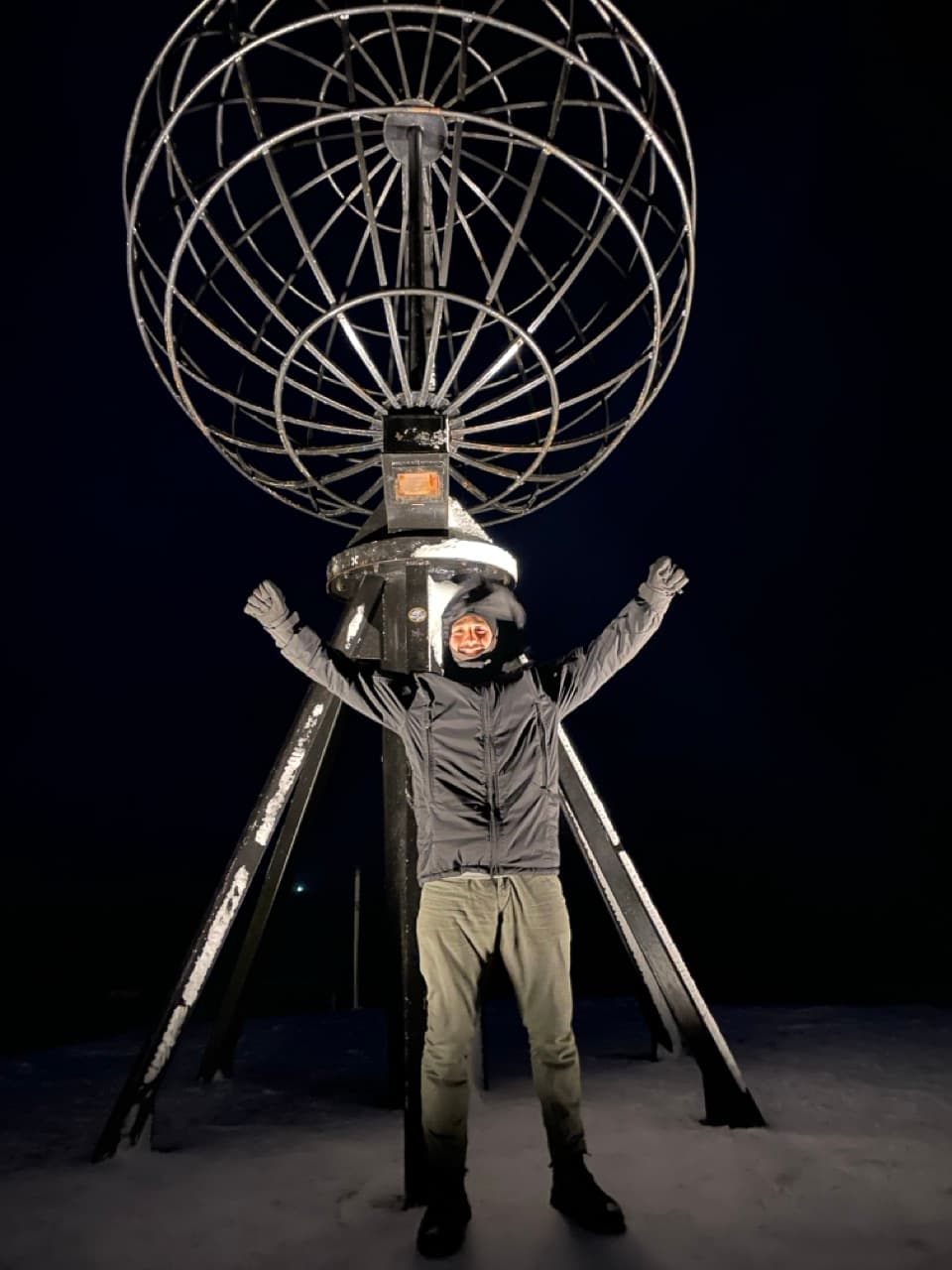 Eine Person in Winterkleidung steht nachts mit erhobenen Armen unter einer großen Metallkugel auf einer verschneiten Fläche.
