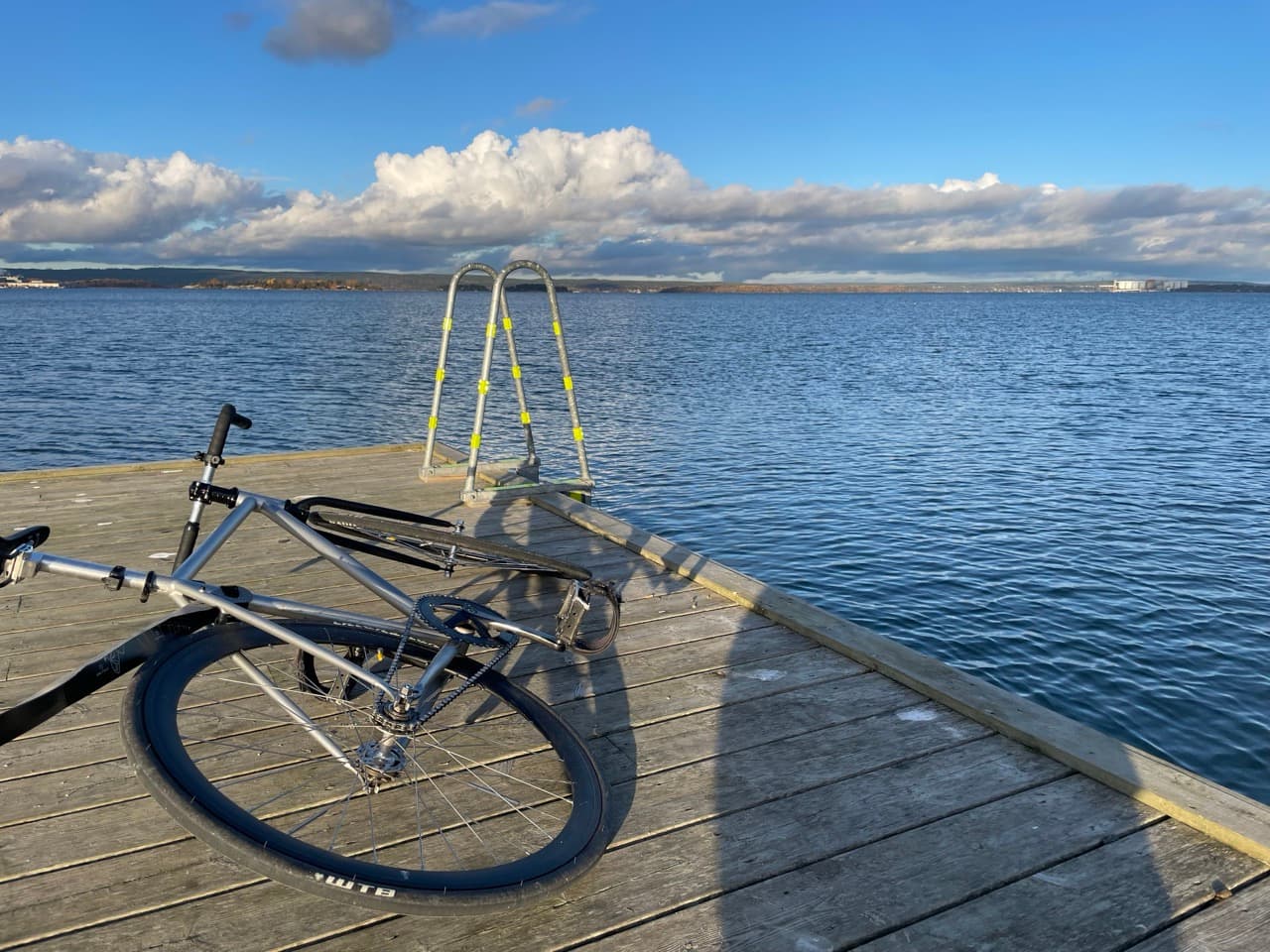 Ein Fahrrad liegt auf der Seite auf einem hölzernen Steg mit Blick auf ein großes Gewässer, mit einer Metallleiter am Rand des Stegs und entfernten Wolken und der Küstenlinie unter einem strahlend blauen Himmel.
