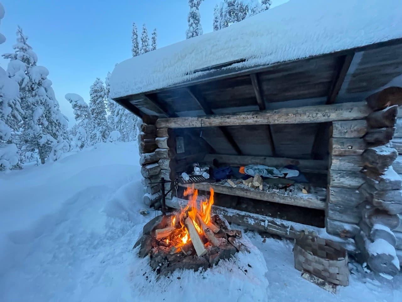 Eine verschneite Blockhütte in einem Winterwald, mit Schlafsäcken und Ausrüstung darin. Davor brennt ein helles Lagerfeuer, umgeben von Schnee und gefrorenen Kiefern.