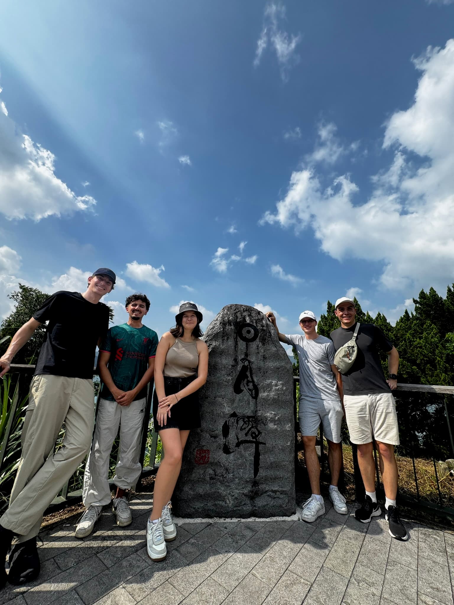 Fünf Menschen stehen im Freien um einen großen Stein mit schwarzen Markierungen und lächeln unter einem strahlend blauen Himmel mit Wolken und Bäumen im Hintergrund.
