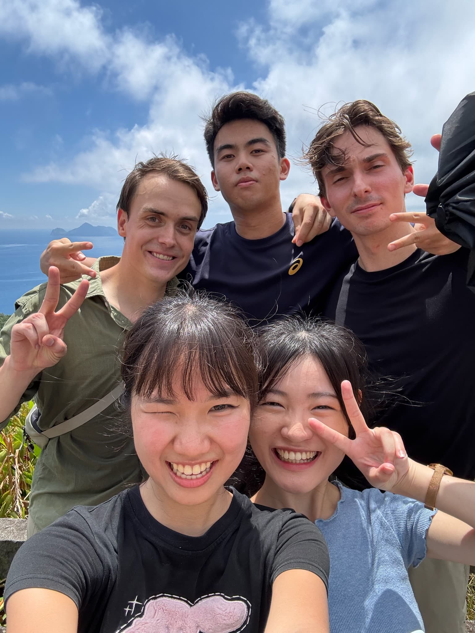 Fünf junge Erwachsene, drei Männer und zwei Frauen, lächeln und zeigen Peace-Zeichen, während sie an einem sonnigen Tag mit blauem Himmel und Meer im Hintergrund für ein Selfie posieren.