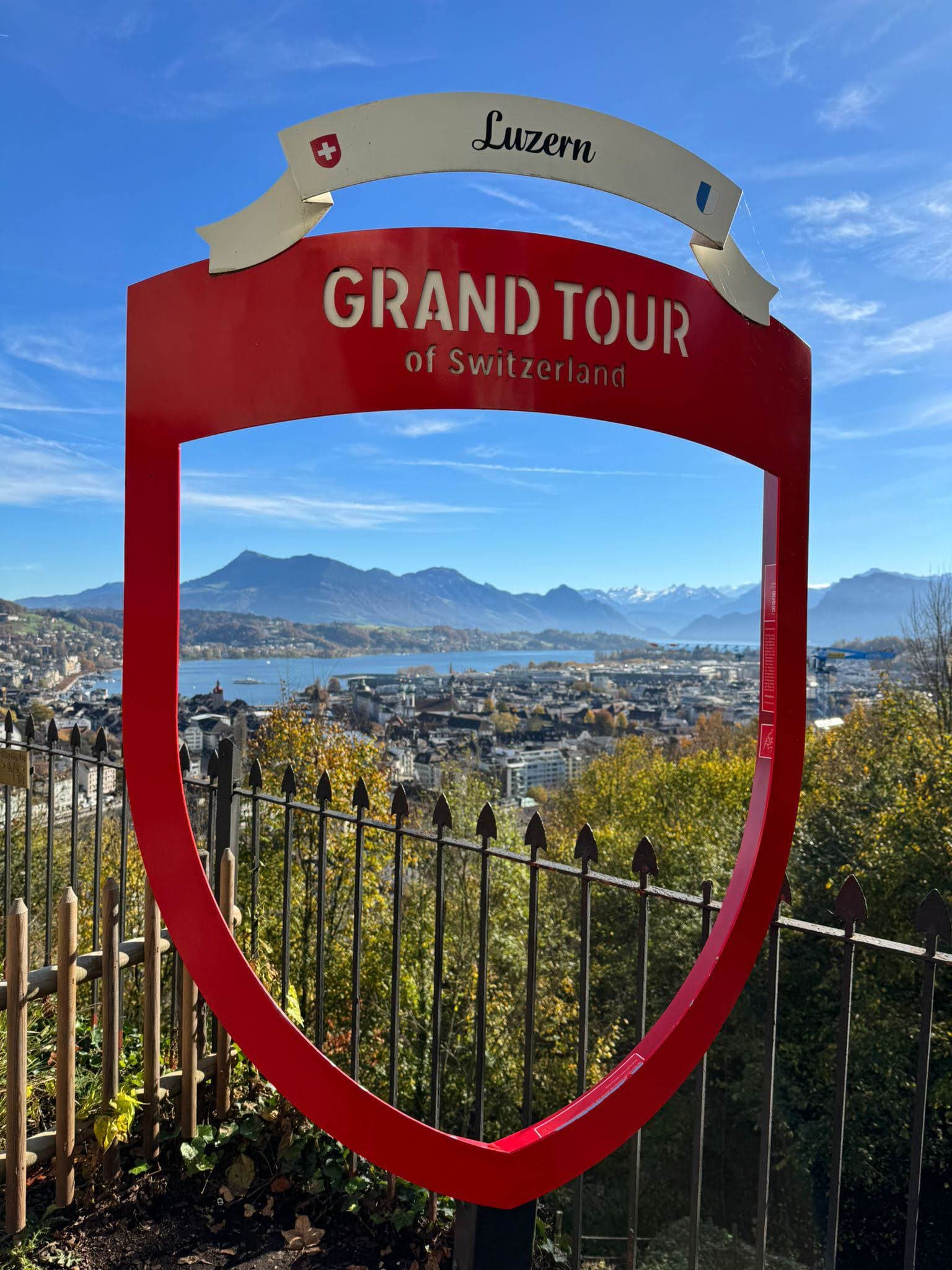 Ein großes rotes Schild mit der Aufschrift Grand Tour of Switzerland umrahmt eine malerische Ansicht von Luzern, mit der Stadt, dem See, den Bergen und dem blauen Himmel im Hintergrund. Im Vordergrund sind Bäume und ein Metallzaun zu sehen.