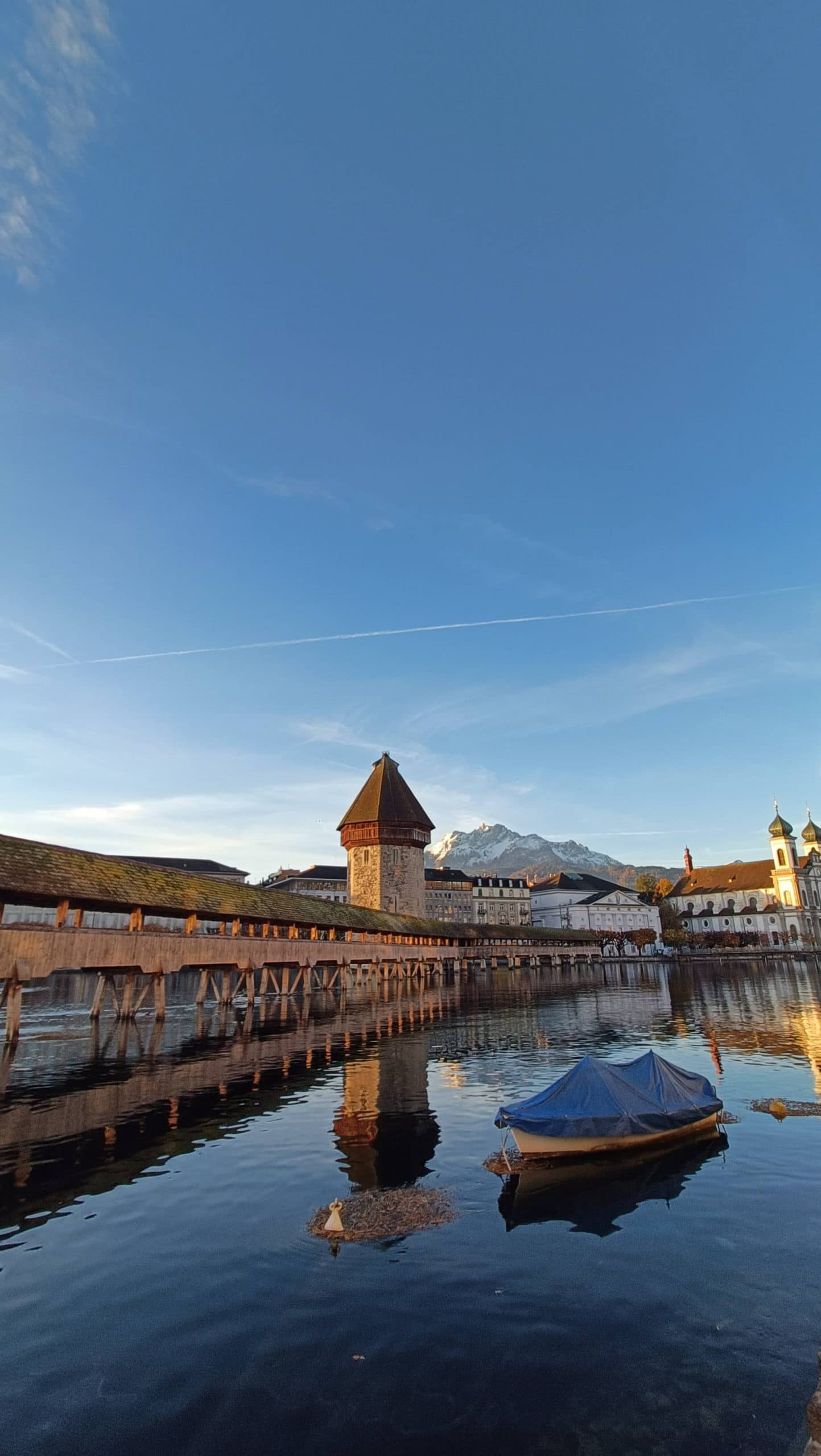 Ein hölzerner Steg und ein Steinturm stehen über dem ruhigen Wasser, in dessen Nähe ein kleines überdachtes Boot schwimmt; im Hintergrund sind Berge und Gebäude unter einem klaren blauen Himmel zu sehen.