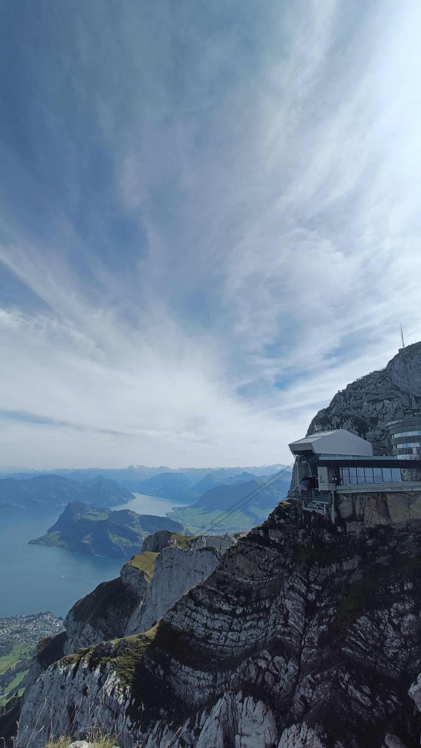 Ein modernes Gebäude steht auf einer felsigen Bergklippe mit Blick auf einen großen blauen See und ferne Berge unter einem dramatischen, teilweise bewölkten Himmel.