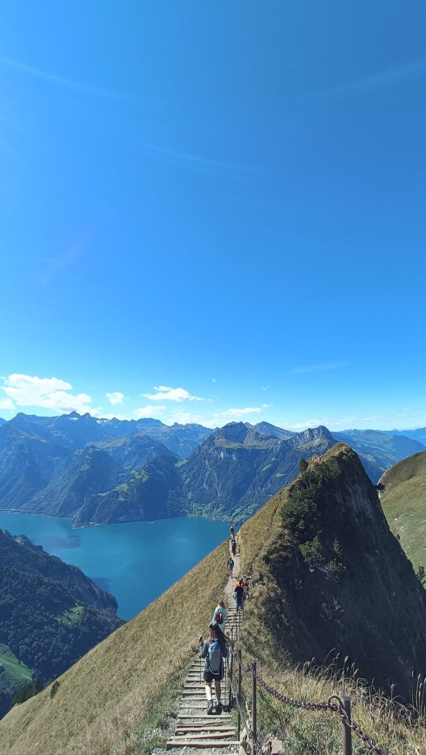 Der Wanderer wandert auf einem schmalen Bergkammweg mit Stufen, mit Blick auf einen blauen See und umgeben von hohen grünen Bergen unter einem klaren, strahlend blauen Himmel.