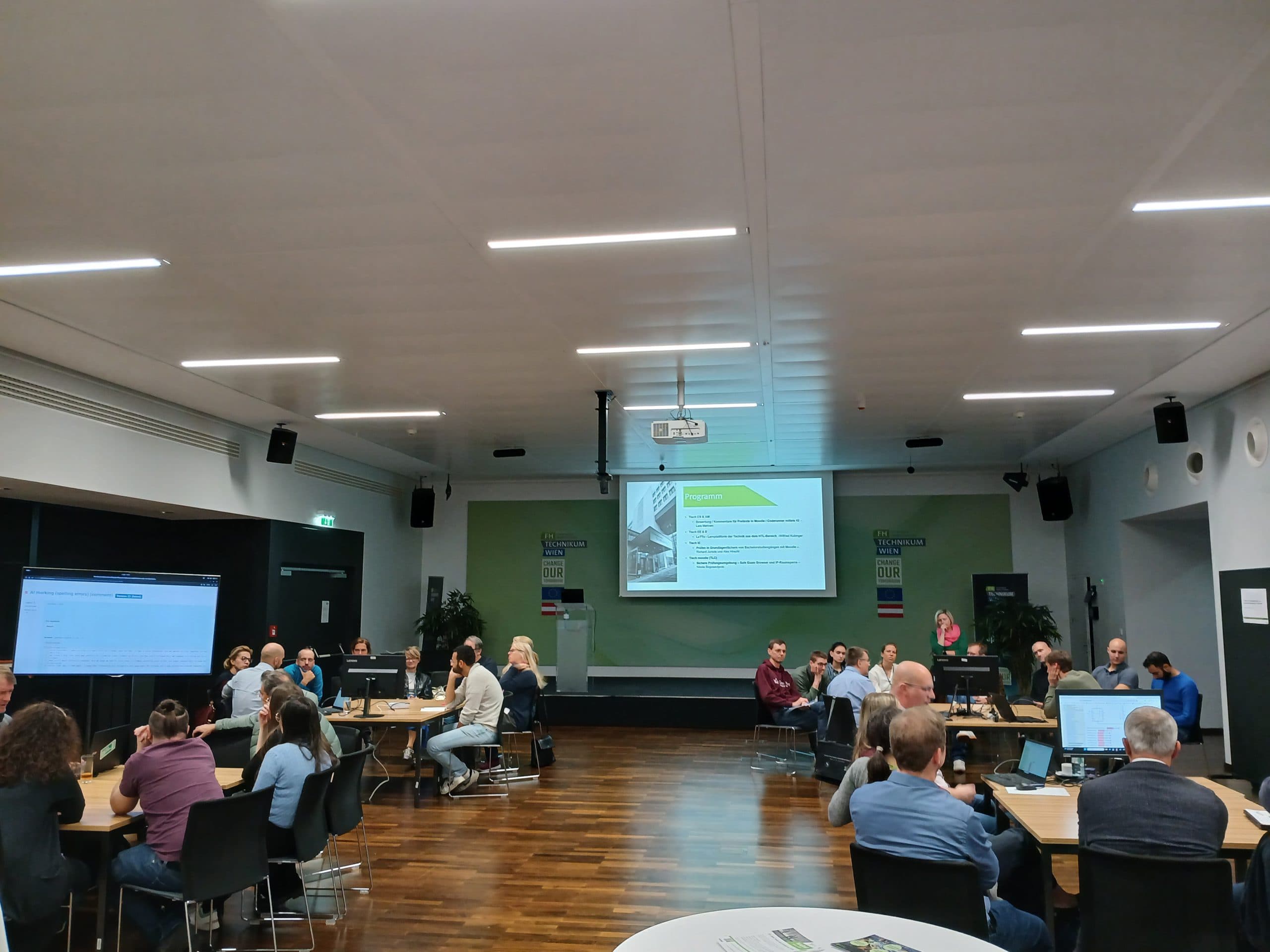 Die Teilnehmer sitzen an runden Tischen in einem gut beleuchteten Konferenzraum. Auf einem großen Bildschirm an der Stirnseite wird eine Präsentation gezeigt, und die Teilnehmer scheinen sich an Diskussionen zu beteiligen oder zuzuhören.